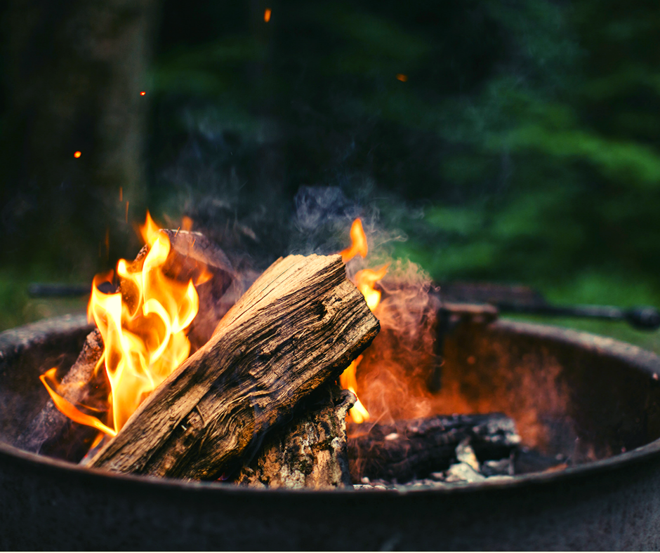 Campfire burning in a metal fire pit with flames and smoke, set against a blurred green backdrop.