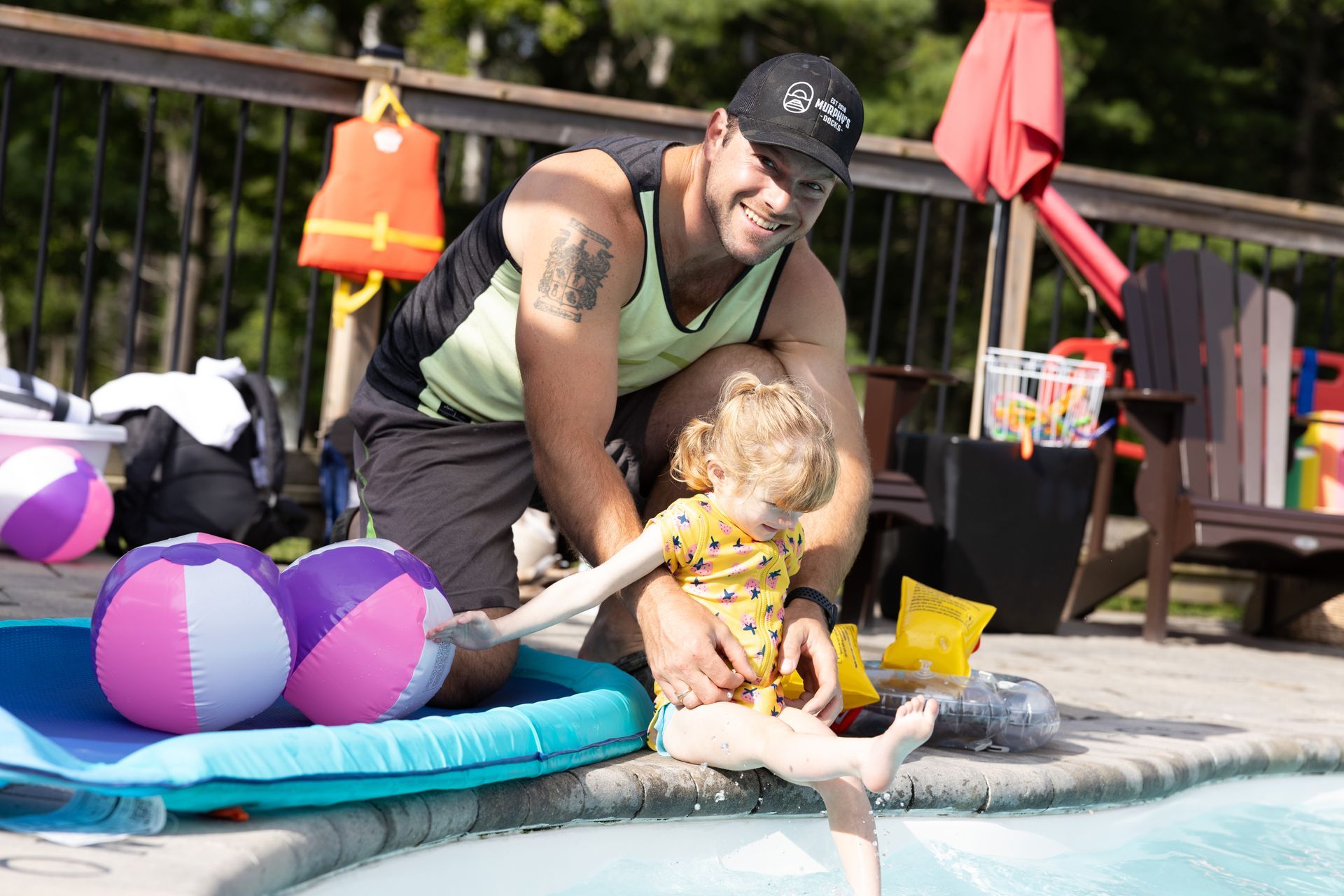 Man kneels beside a child at a pool's edge. Both smile. Two beach balls and pool toys sit nearby on a blue float.