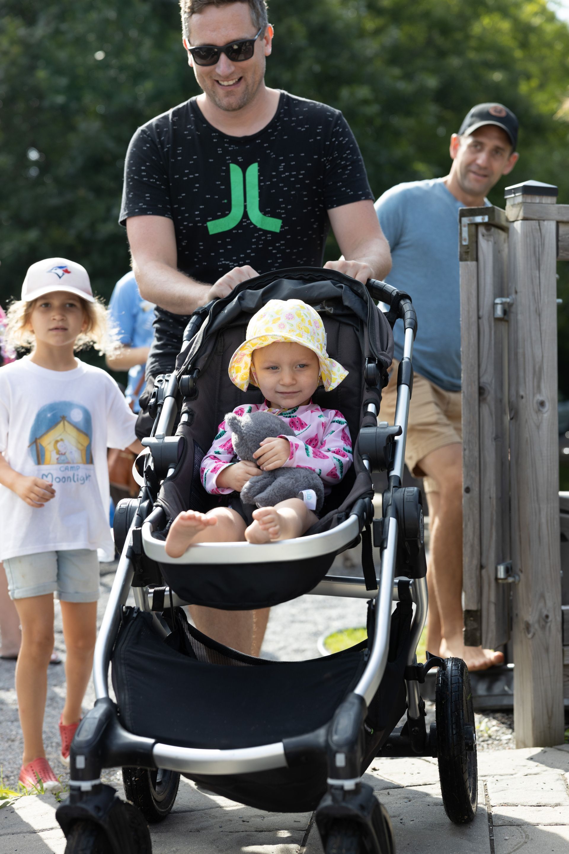 Family pushing stroller with child; park setting, sunny day.