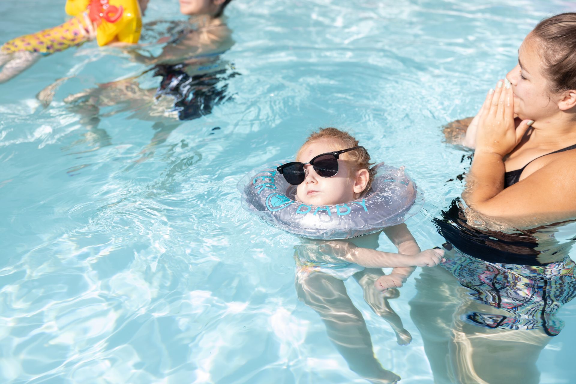 Baby in a swim float with sunglasses in a pool, supported by an adult.