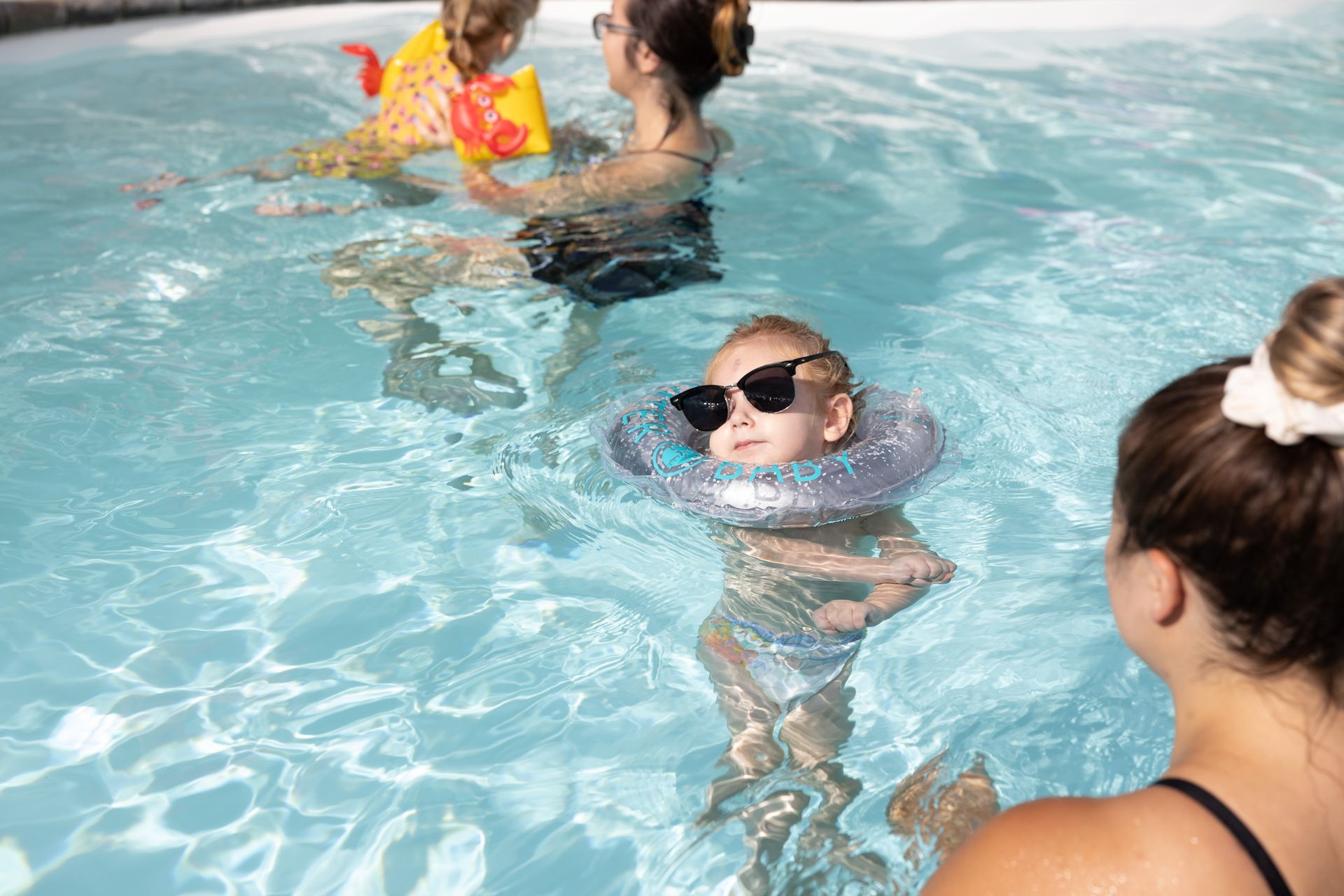 Child wearing sunglasses and a neck float in a pool with adults and another child playing nearby.