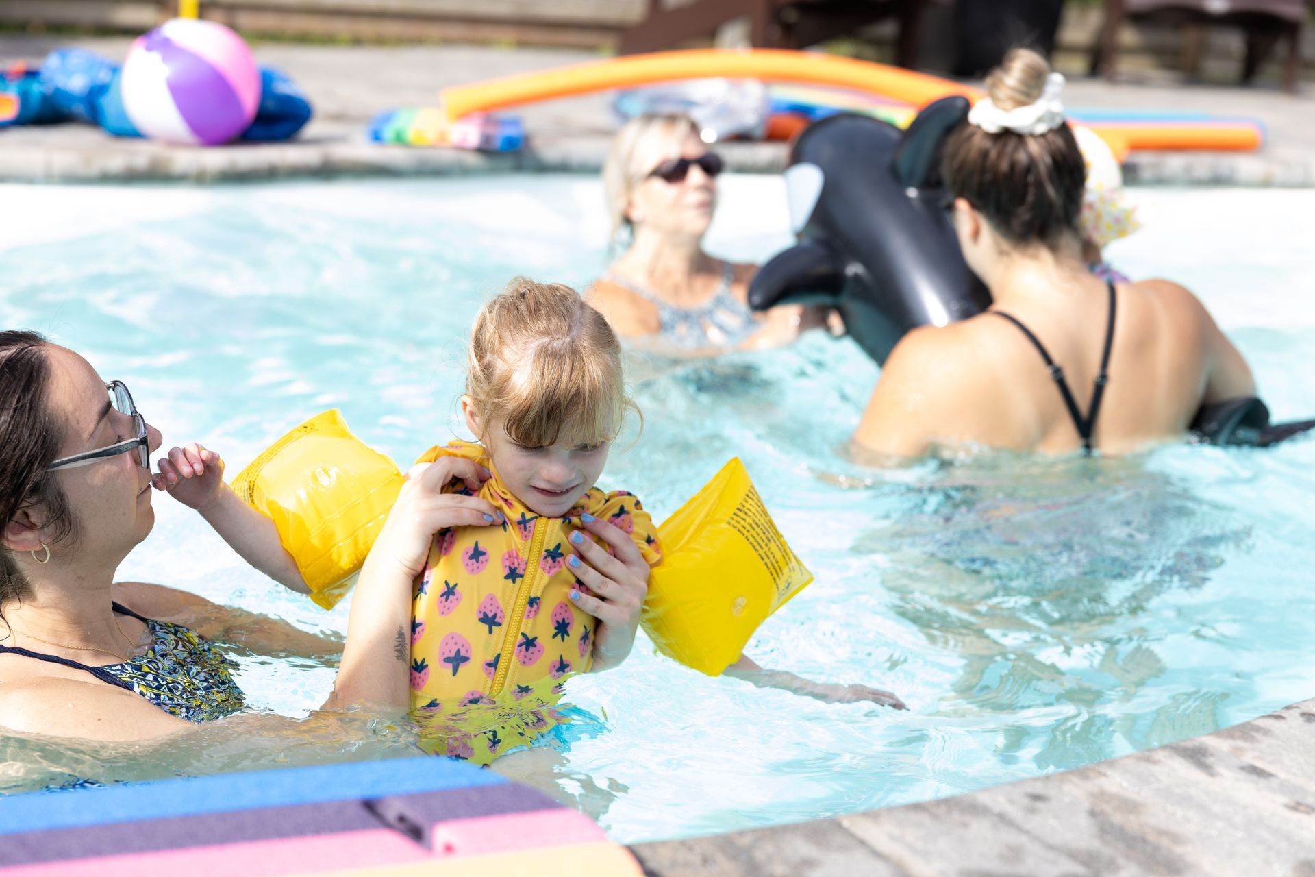 A child with arm floats is held in a pool by an adult. Other people swim nearby with inflatable toys.