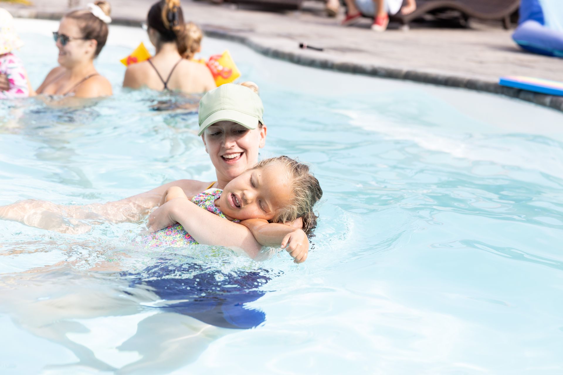 Woman in a pool holding a smiling child, both enjoying the water. Other people and a pool's edge are visible.