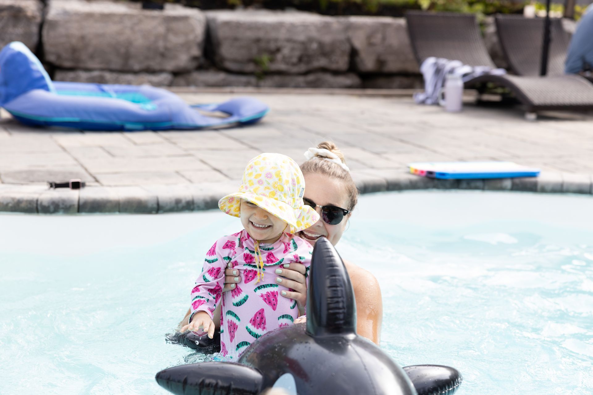 Woman holding a child in a pool. Child wears a hat and swim shirt. Pool setting with floats.