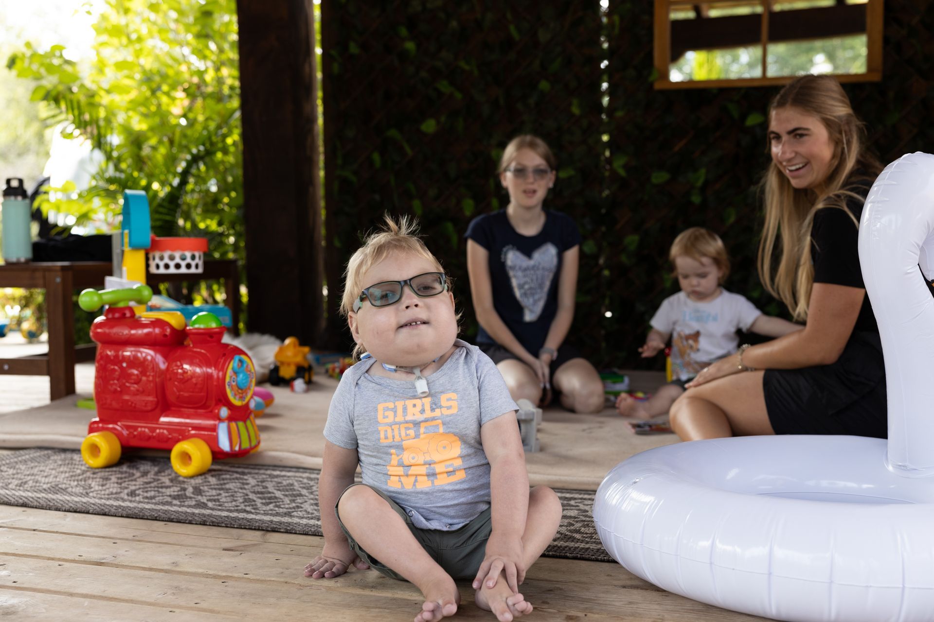 Child wearing sunglasses sits on porch; family members behind him, toys nearby.