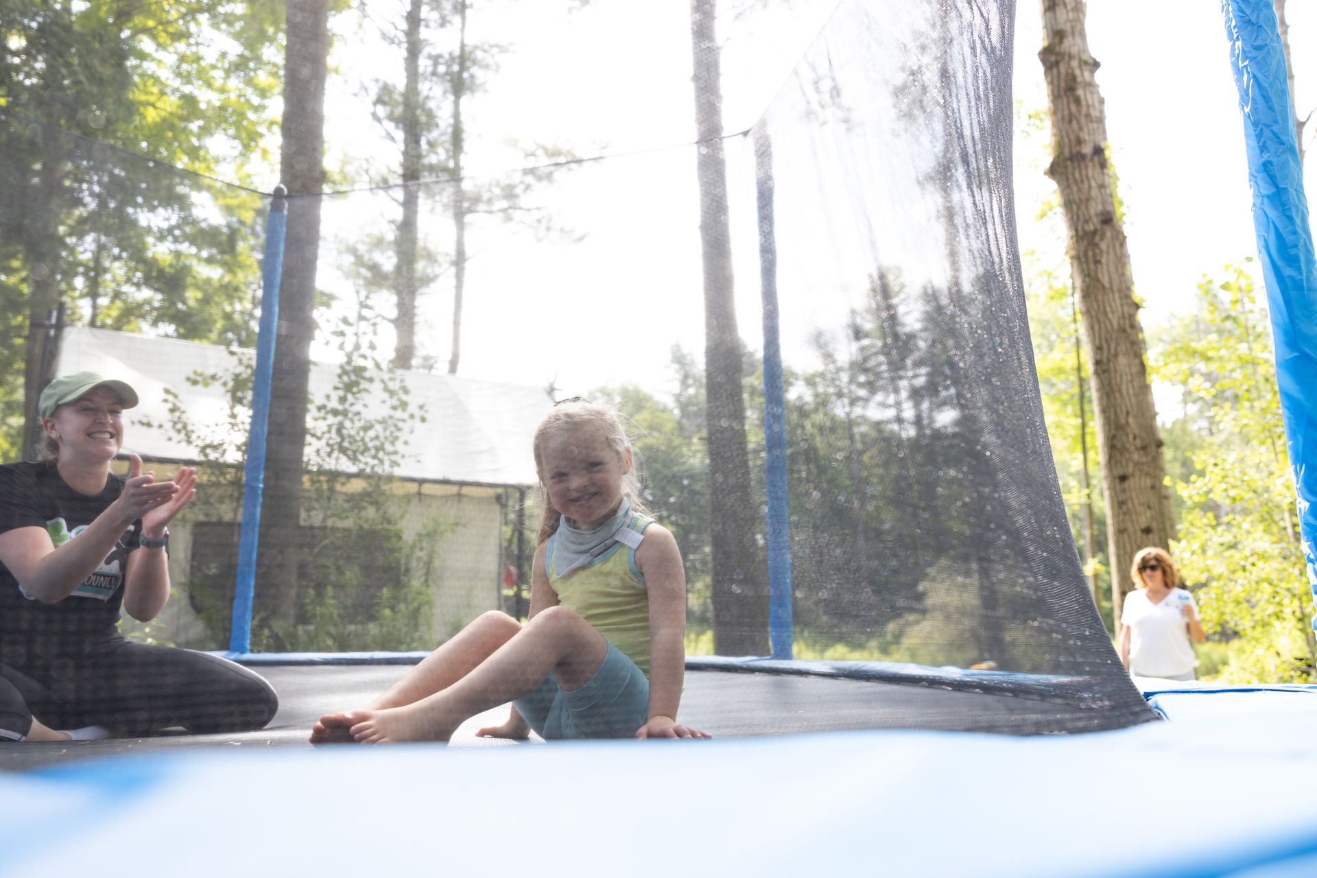 Girl on trampoline smiles, wet from sprinkler. Another person claps nearby in outdoor setting with trees.