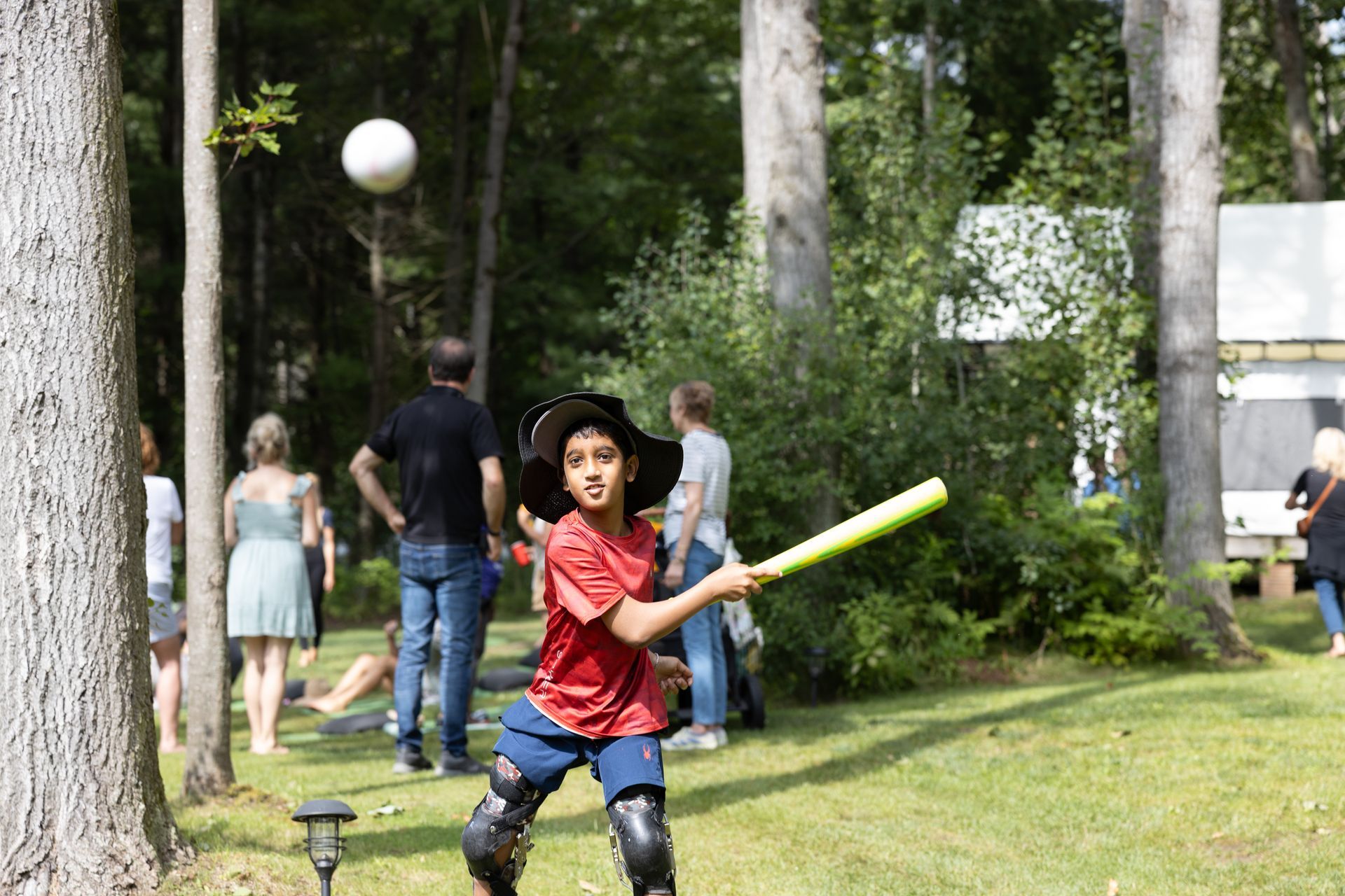 Boy in a hat swings a bat at a ball, with trees and people in a park-like setting.