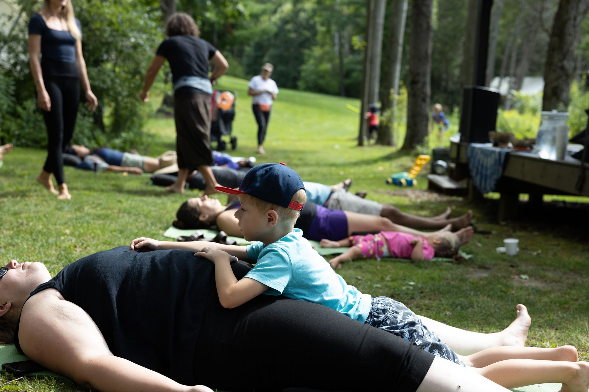 People lying on grass; a child sits on a person’s stomach. Others walk nearby. Outdoors, sunny day.