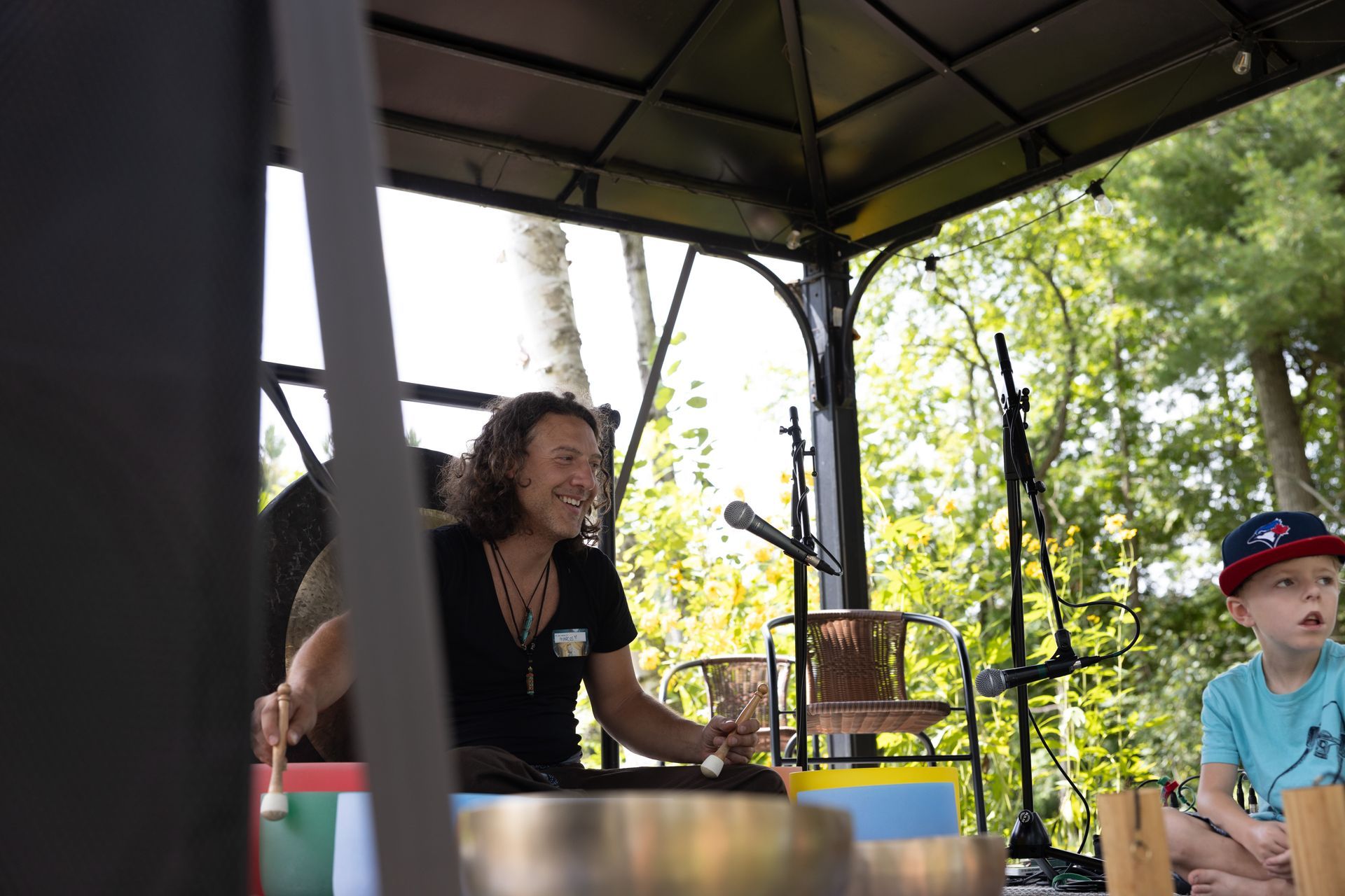 Man playing singing bowls under a gazebo, boy watching. Outdoors, sunny.