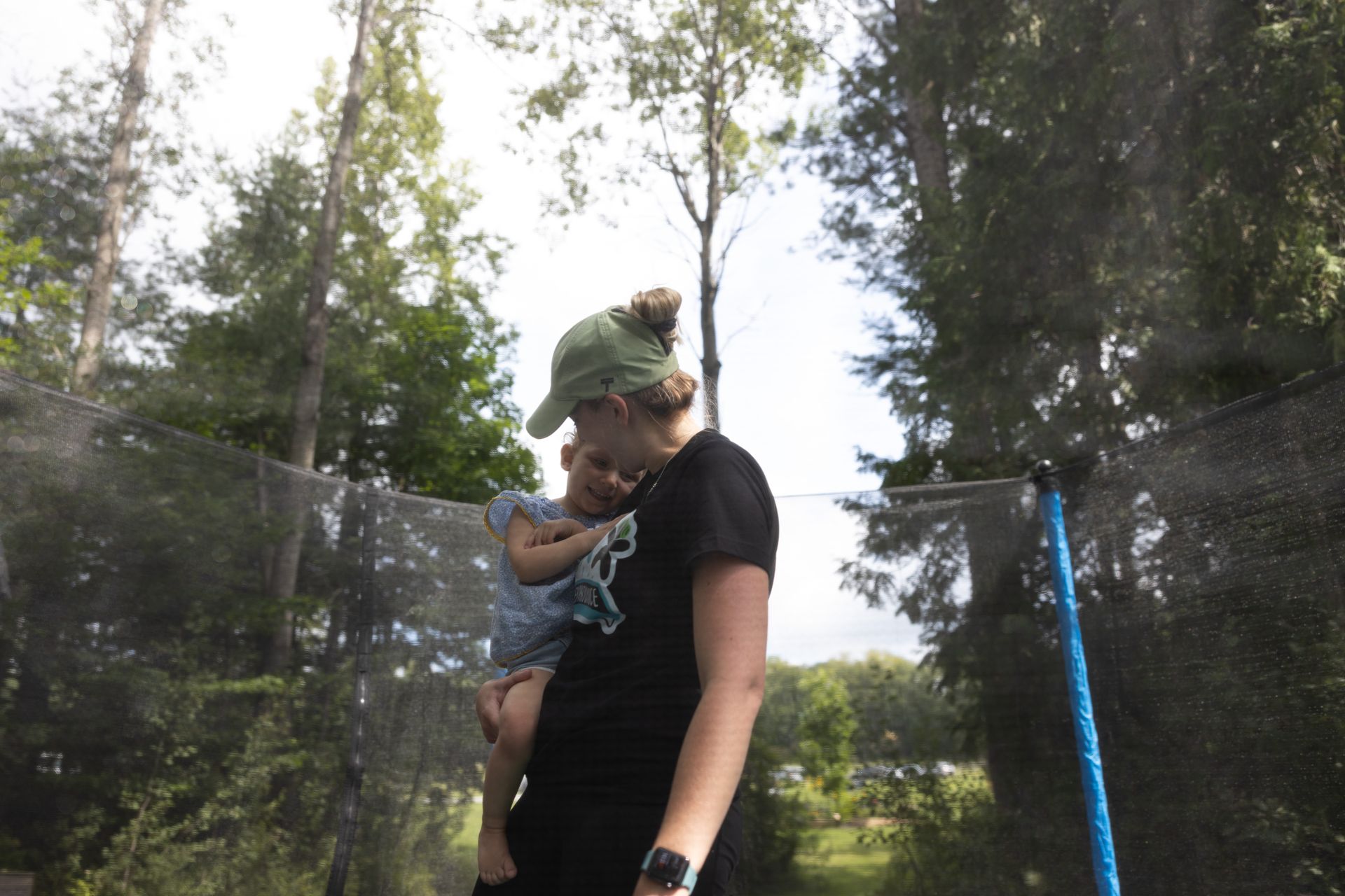 Woman holding child on a trampoline, trees in background, sunny day.