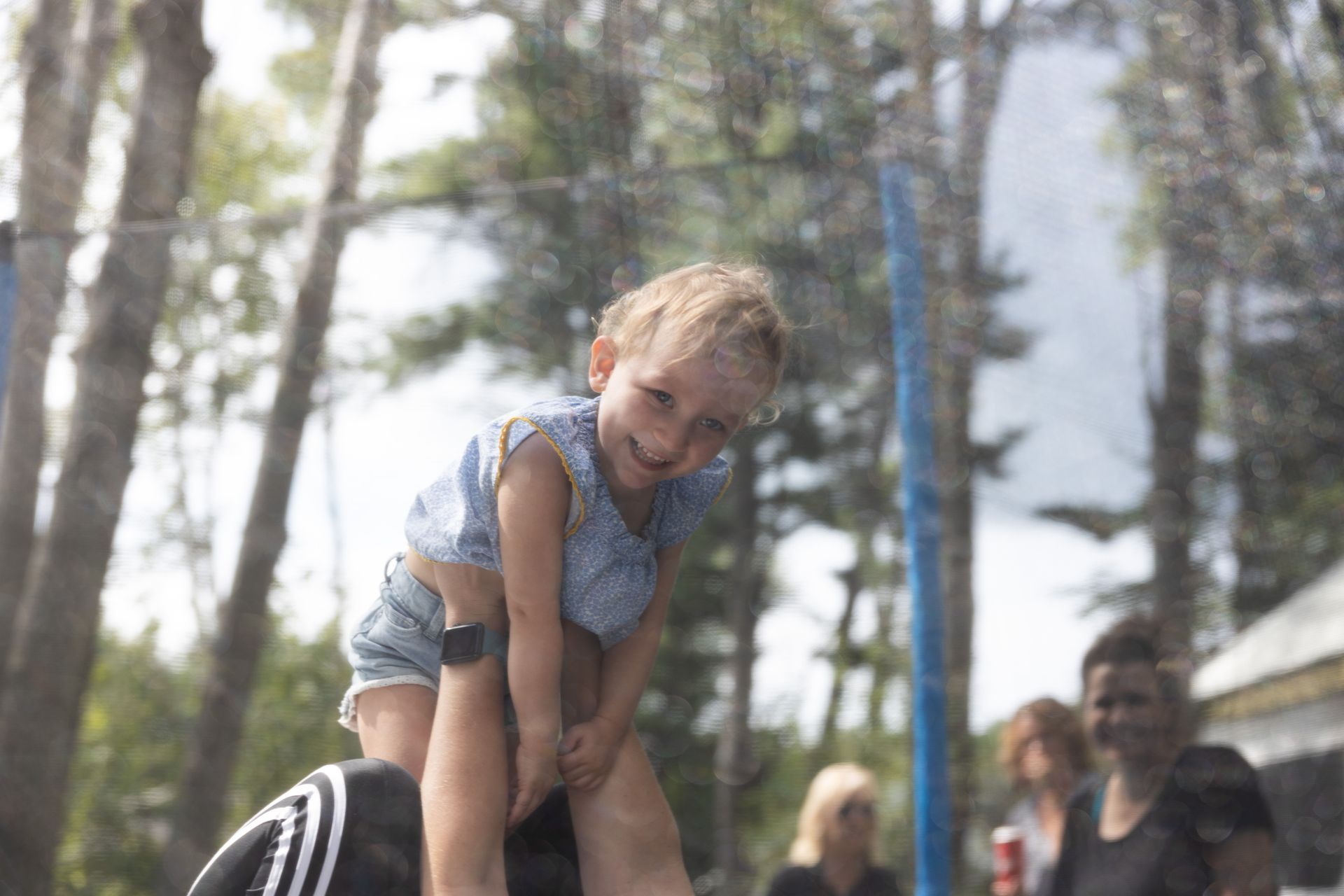 Girl held up, smiling, near trampoline netting, surrounded by trees and people.