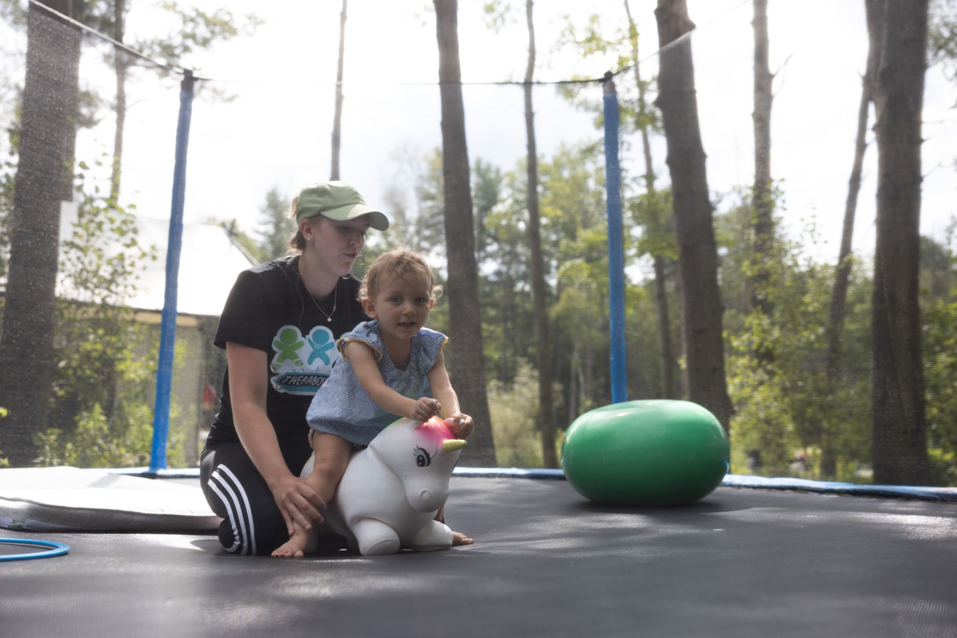 Woman and toddler on a trampoline, toddler bounces on a white inflatable unicorn. Forest setting.