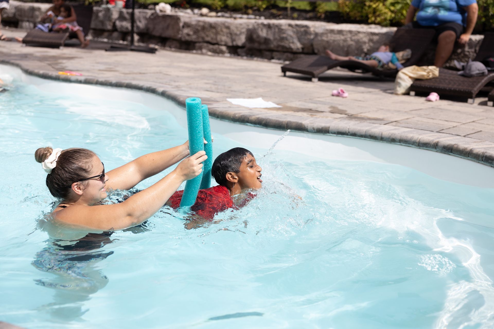 Woman assists boy in pool, using a foam noodle for support.