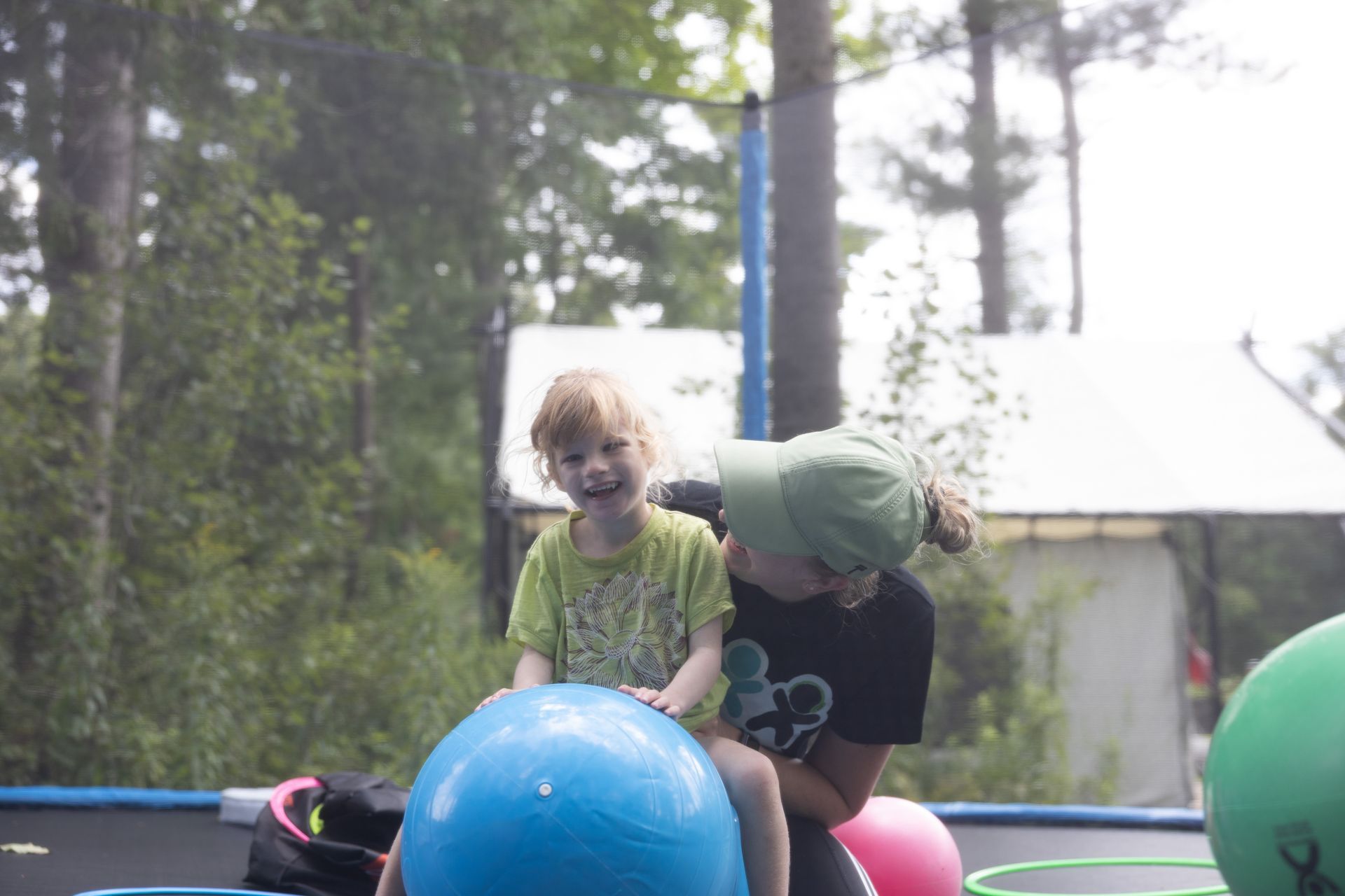 Child and person on a trampoline with a large blue ball, surrounded by trees.