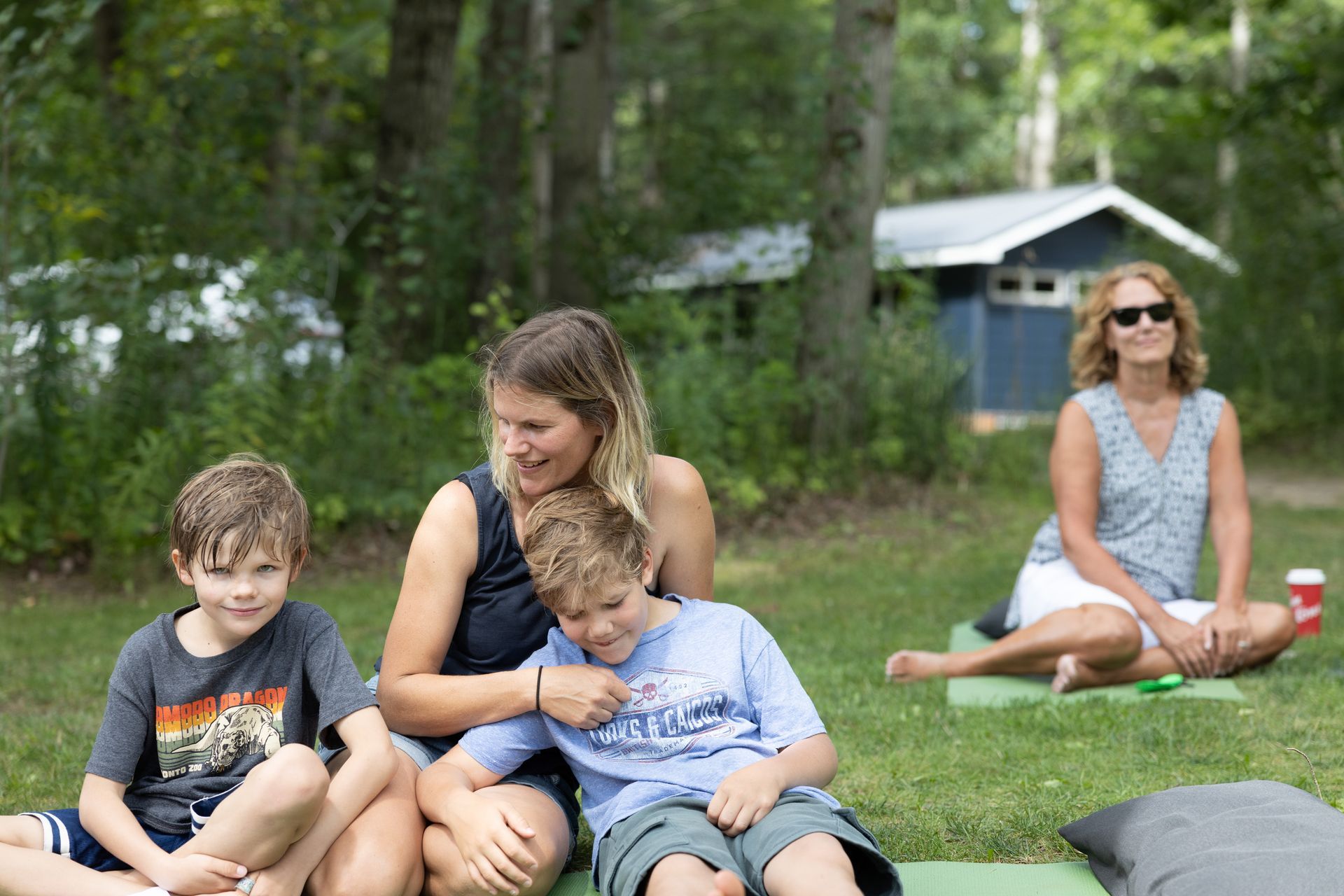 People relaxing on grass. Woman with two children. Another woman sitting on a yoga mat. Blue shed in background.