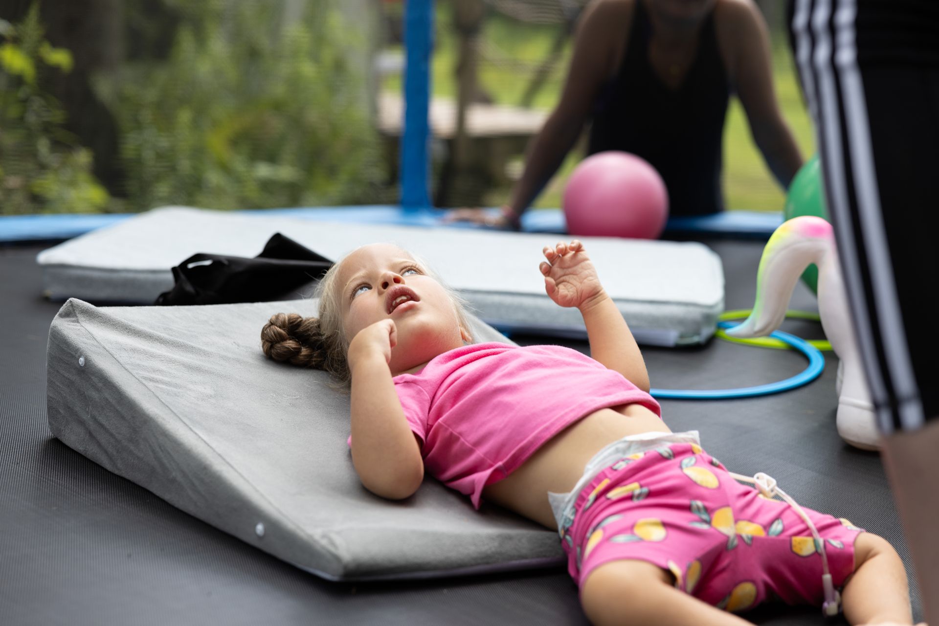 Young child wearing pink shirt and shorts lies on a gray wedge pillow on a trampoline, looking upward.