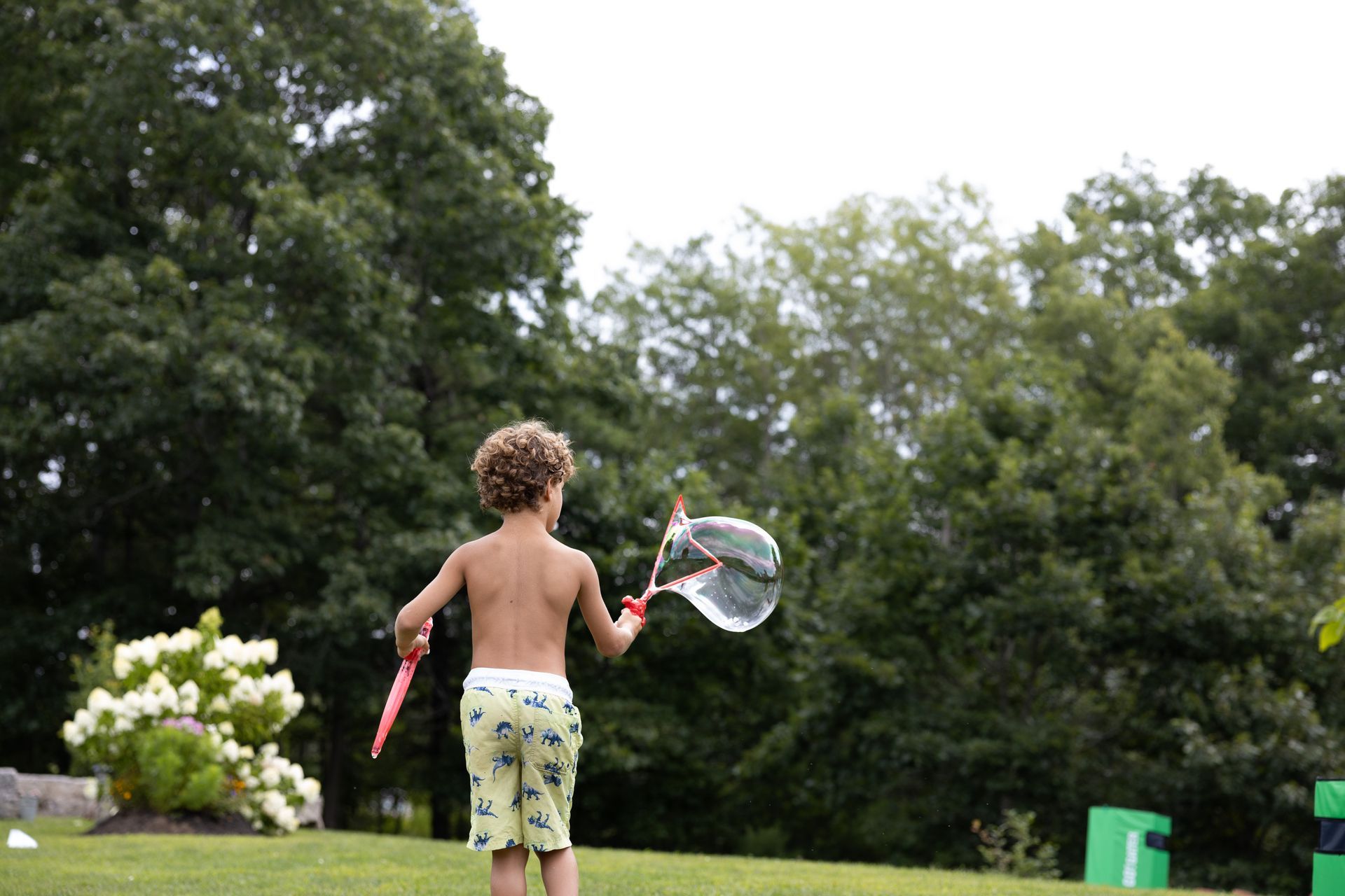 Boy blowing a large bubble in a green yard with trees. He holds the wand and is partially shirtless.