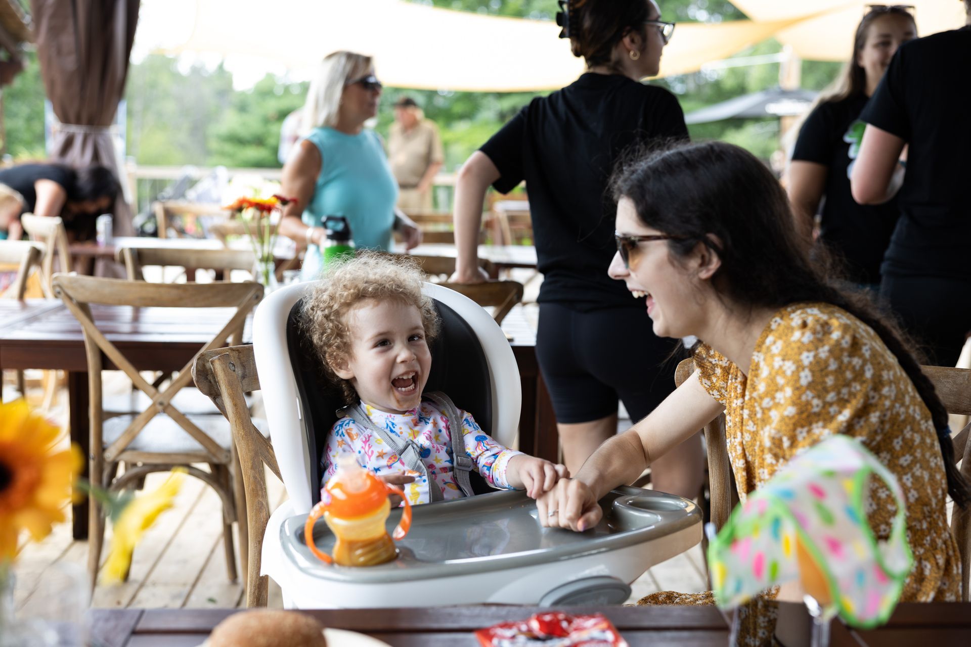 Woman and child laughing at a table. Child in high chair, orange sippy cup, outside setting.