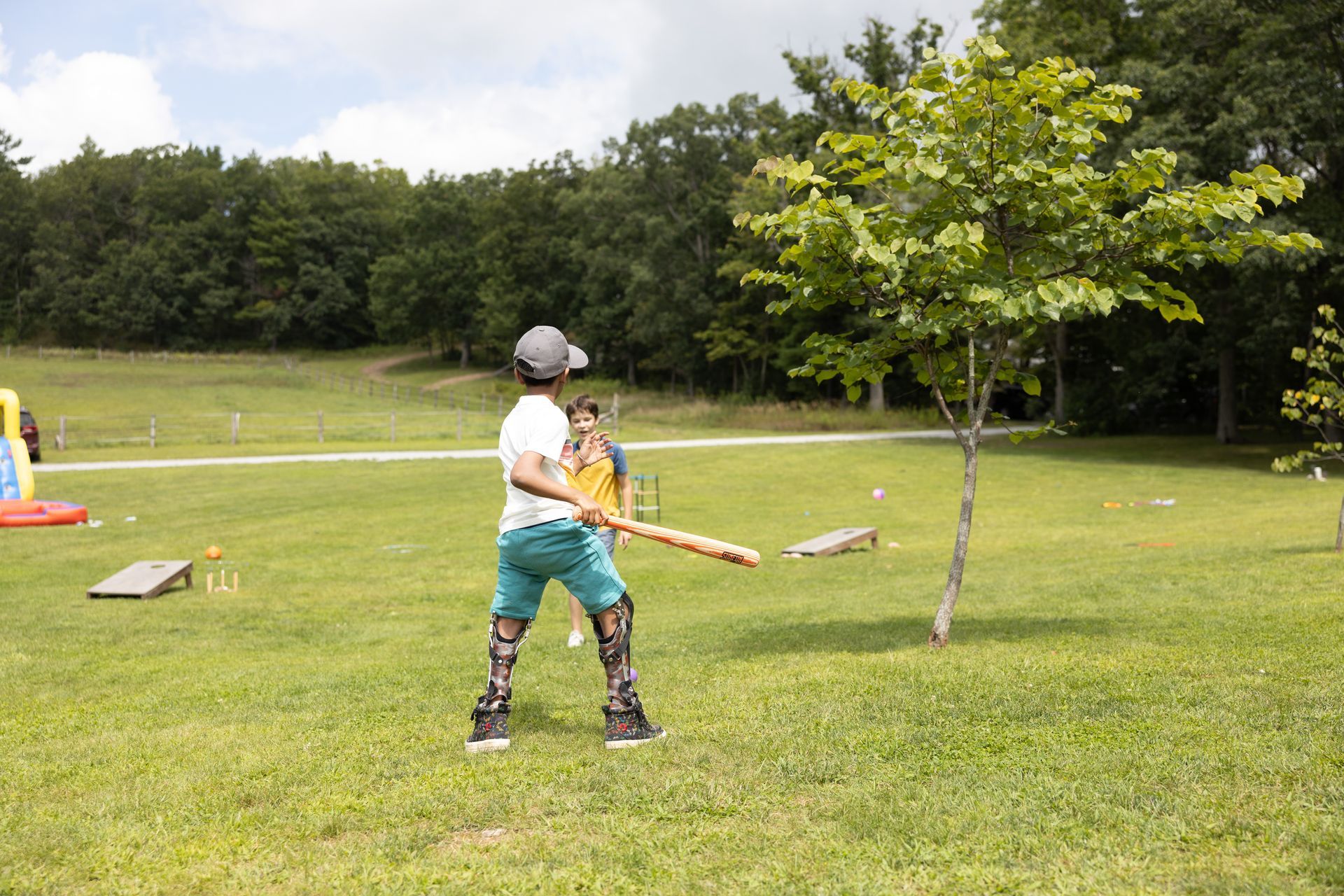 Boy in leg braces swings a bat in a grassy field, trees in the background.