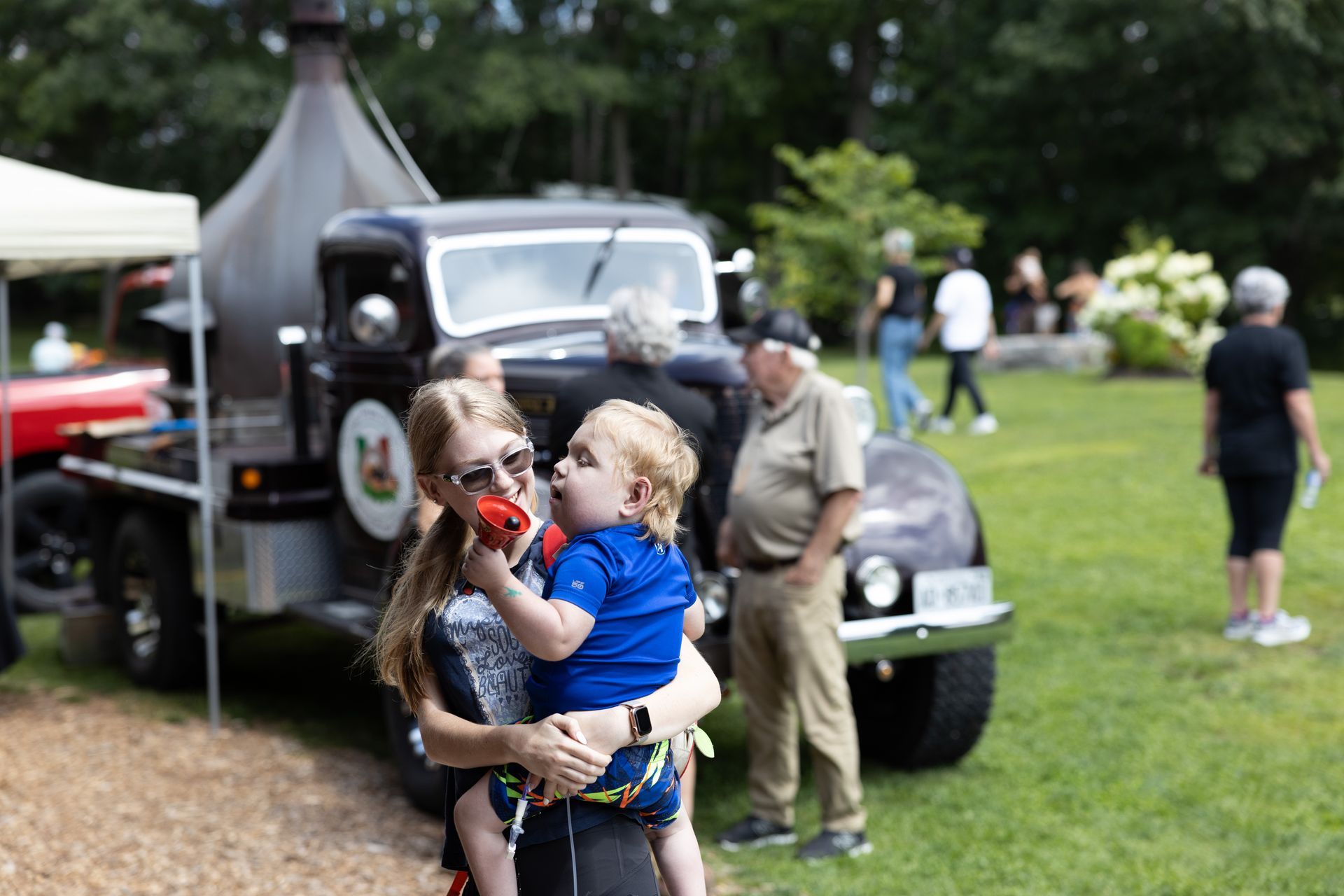 Woman holding child near vintage truck at outdoor event.