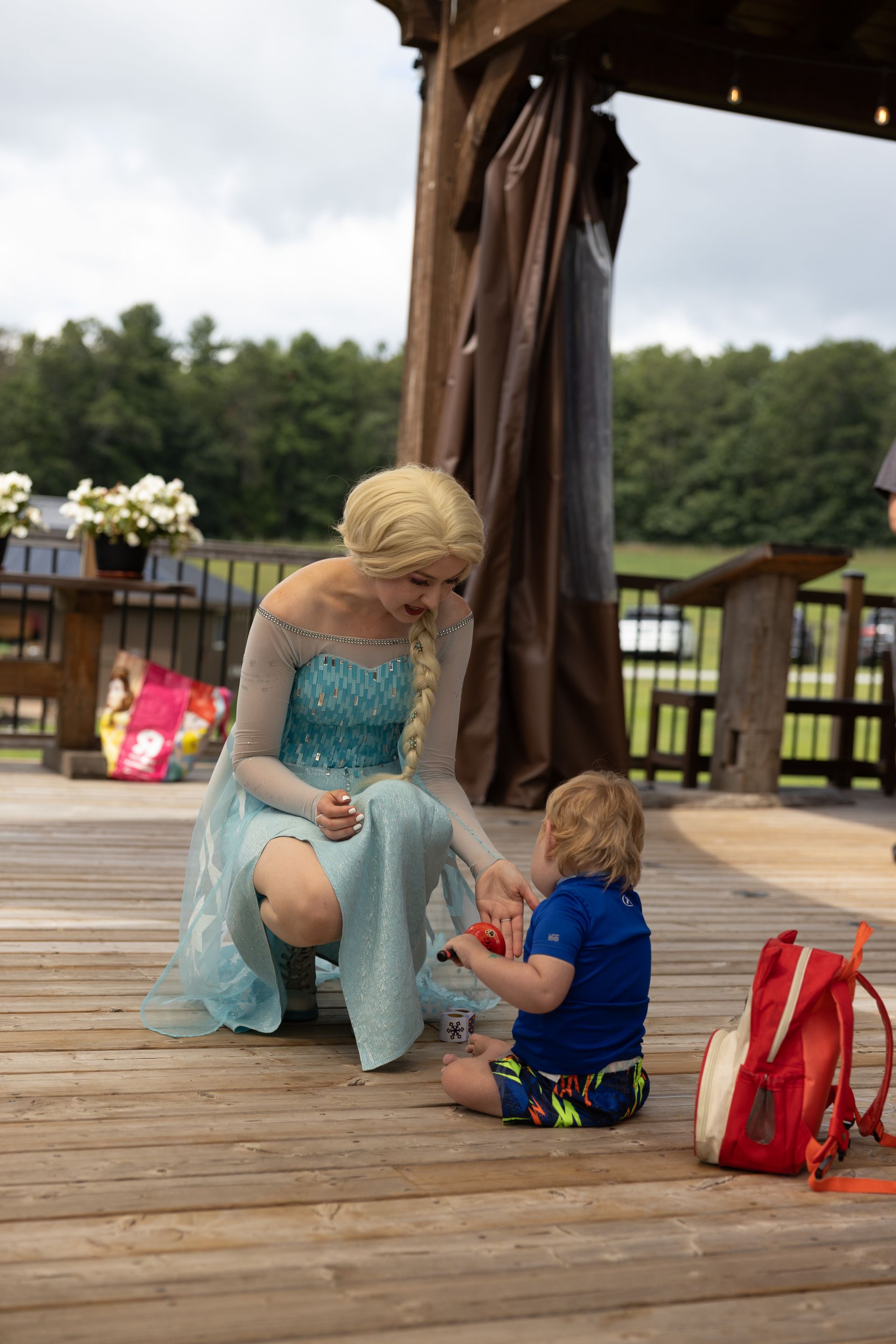 Woman in blue dress kneels to interact with a child on a wooden deck. A red backpack is nearby.