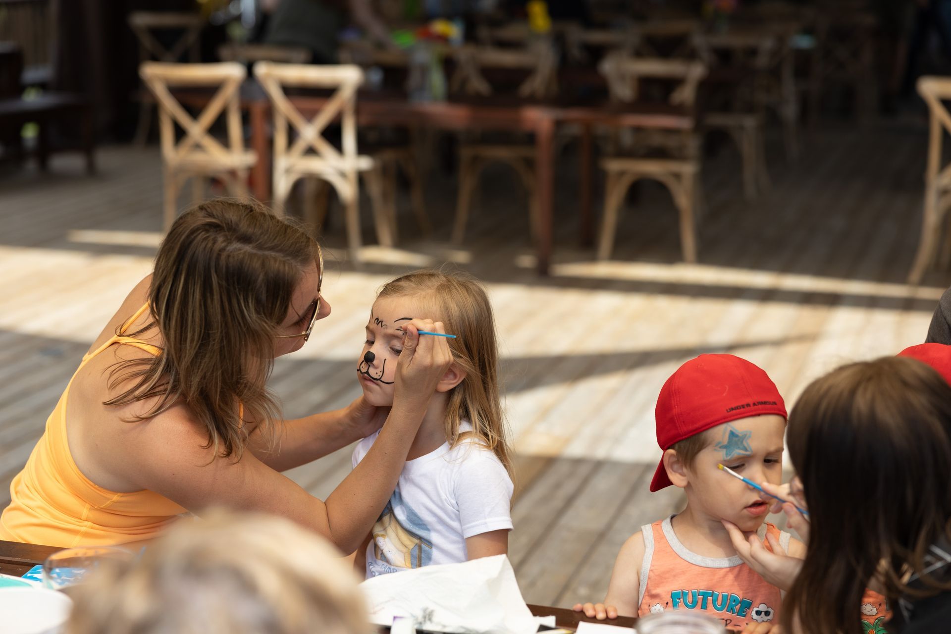 Woman paints a design on a child's face outdoors. Another child has face paint. Tables and chairs in the background.