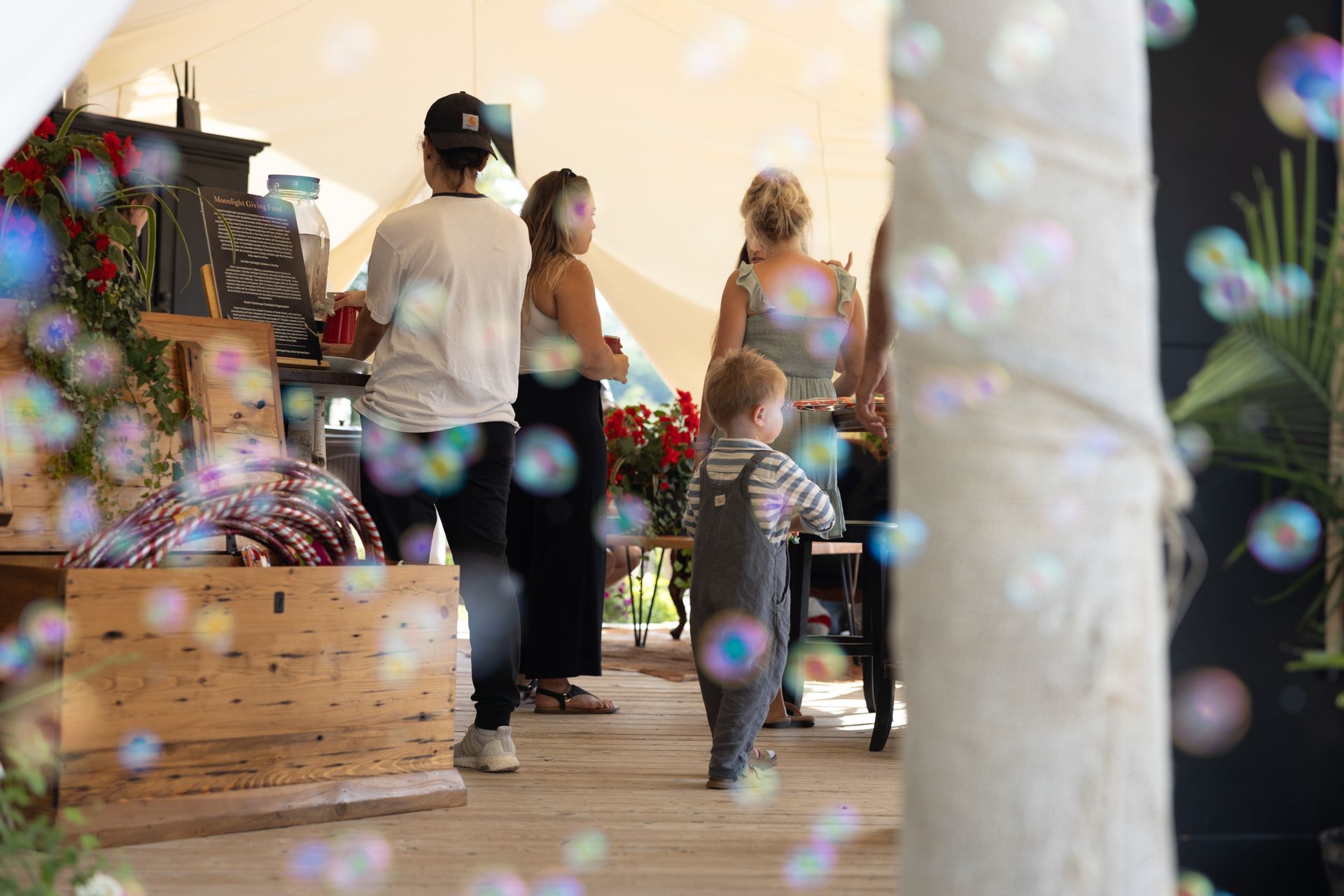 Bubbles float in foreground as people browse an outdoor market; child in overalls watches.
