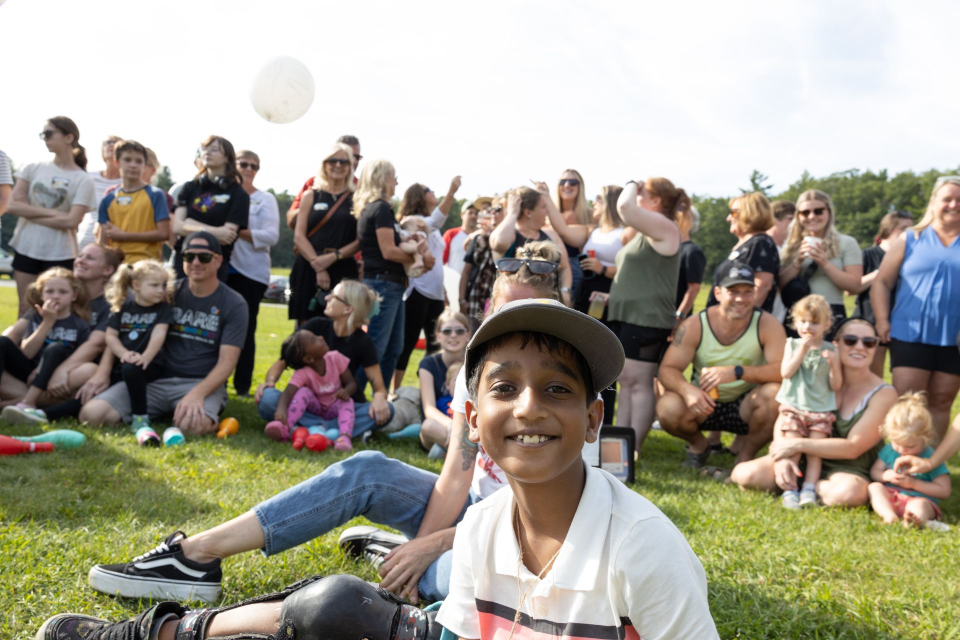 Boy smiling in front of a crowd on a grassy field; white balloon in the air.