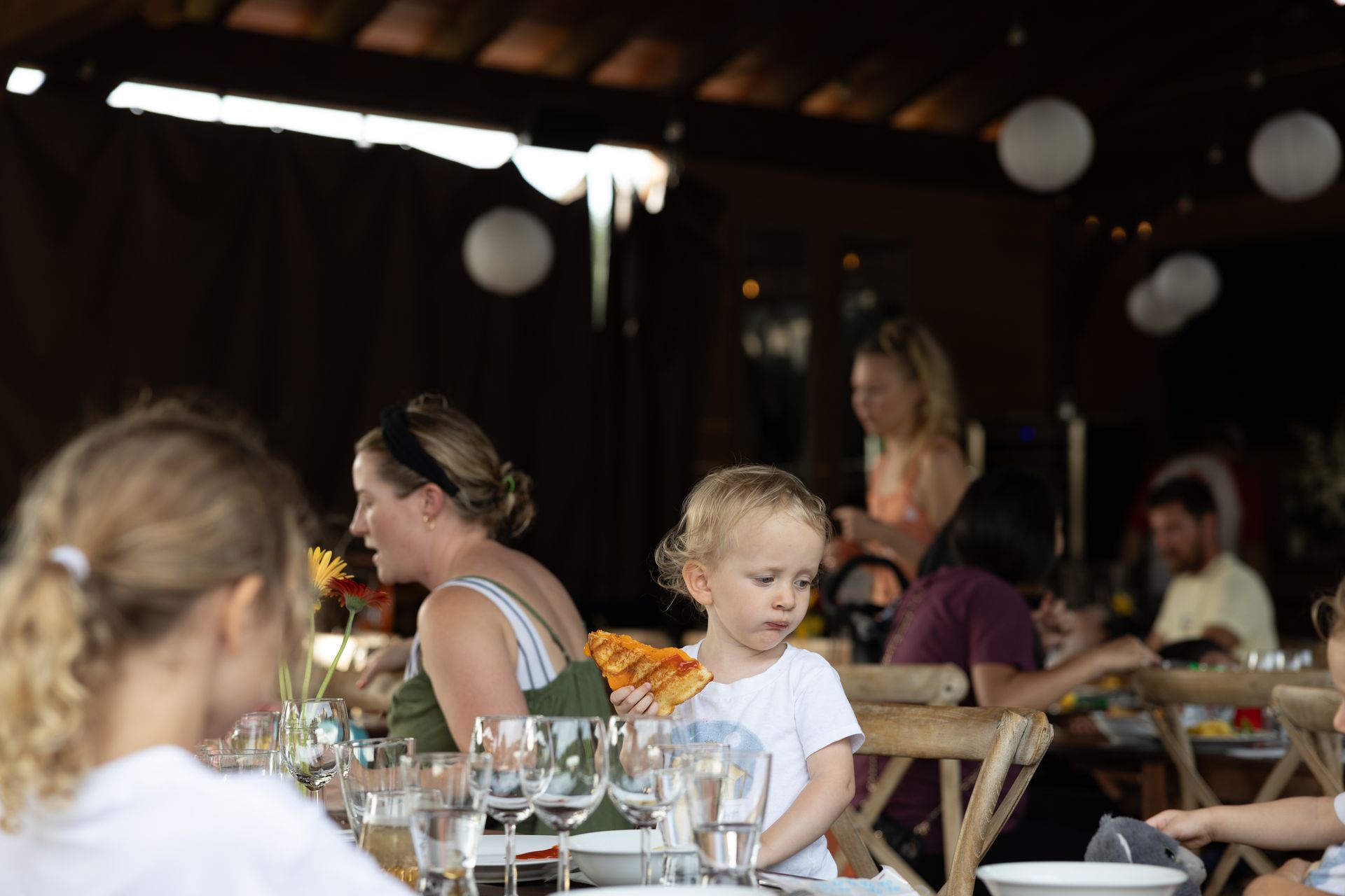 People seated at a dining table under a covered outdoor space; a young child looks down.