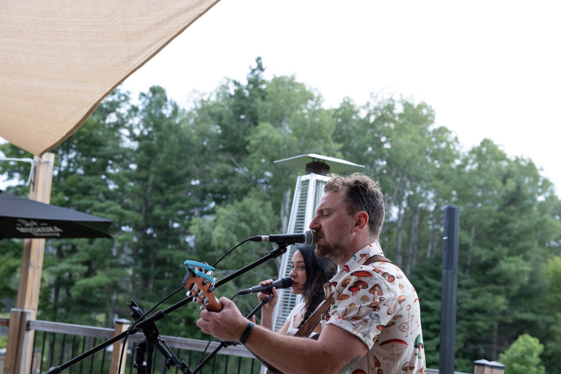 A musical duo performs outdoors: a person plays a ukulele while another sings into a microphone.