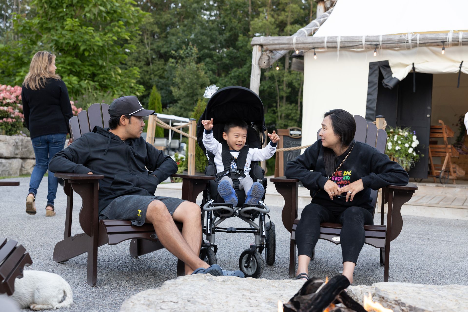 Family sits by a fire pit, a child in a wheelchair raises arms. Outdoor setting with greenery.