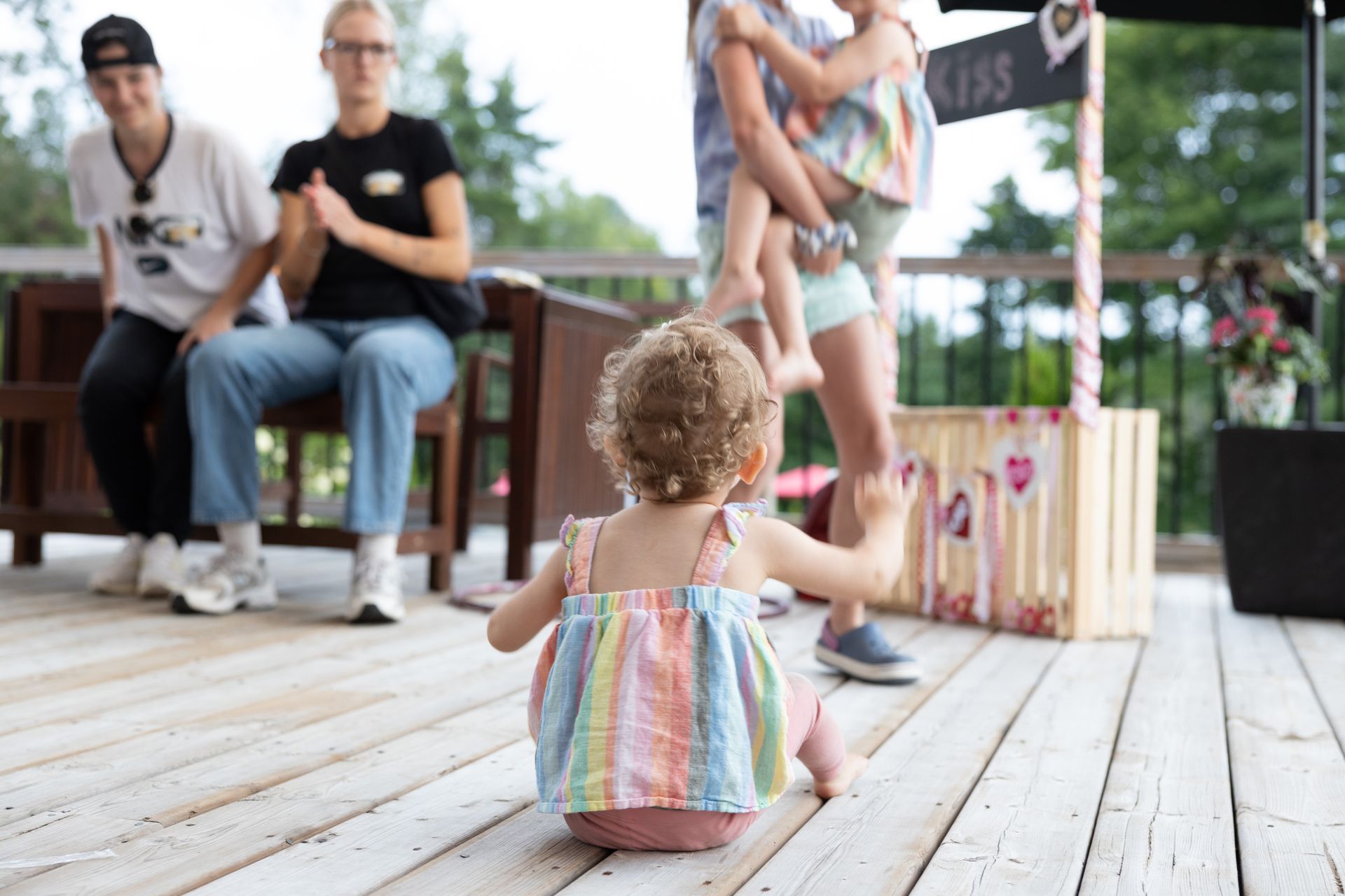 Child on deck reaching towards person holding another child near a lemonade stand.