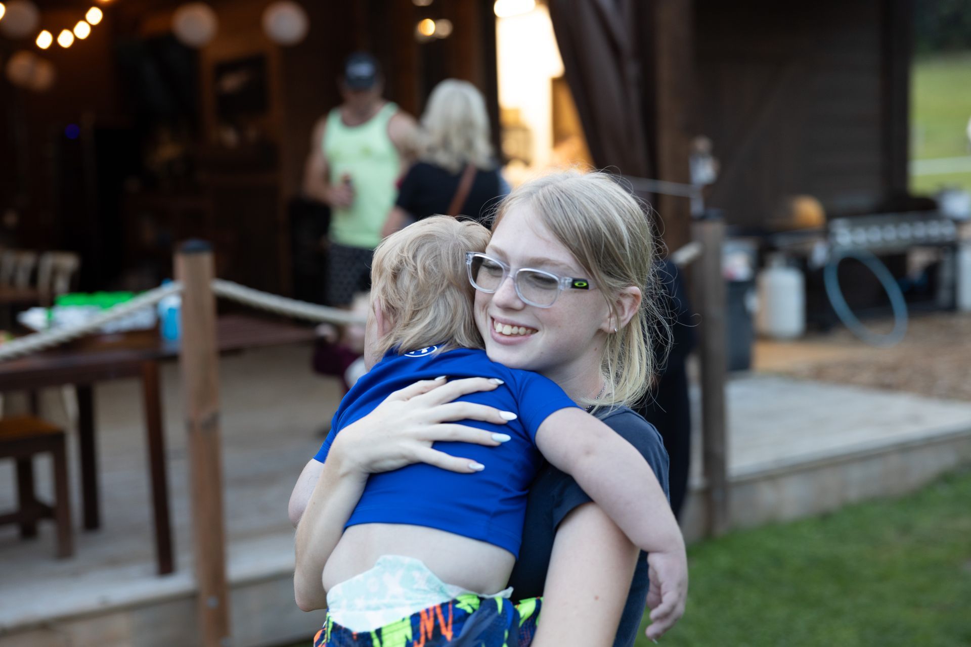 Teenager wearing glasses hugs a young child outside near a building; smiling.