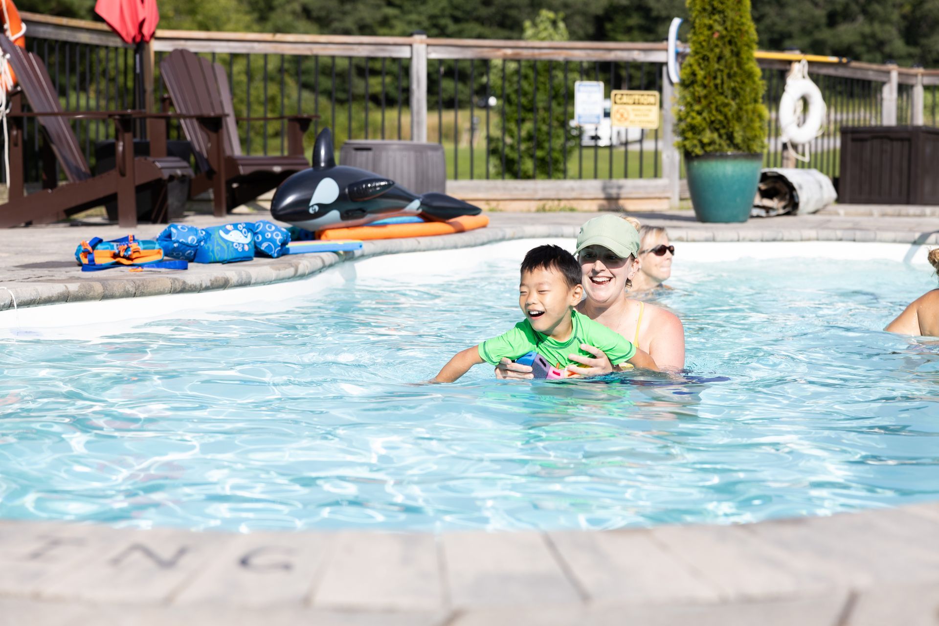 Boy in green shirt held by person in pool, smiling. Other people, pool toys, and wooden deck visible.