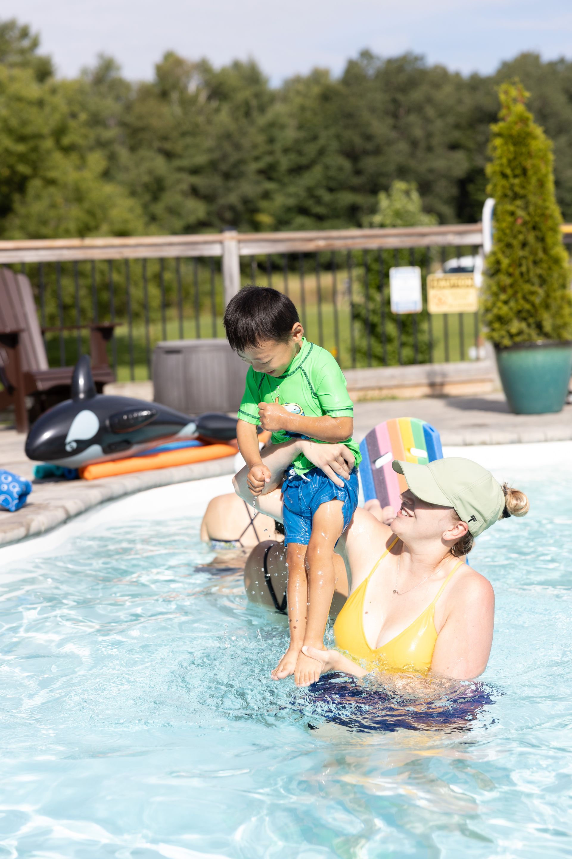 Woman in yellow swimsuit supports a child in a pool. Child wears a green shirt and blue shorts.