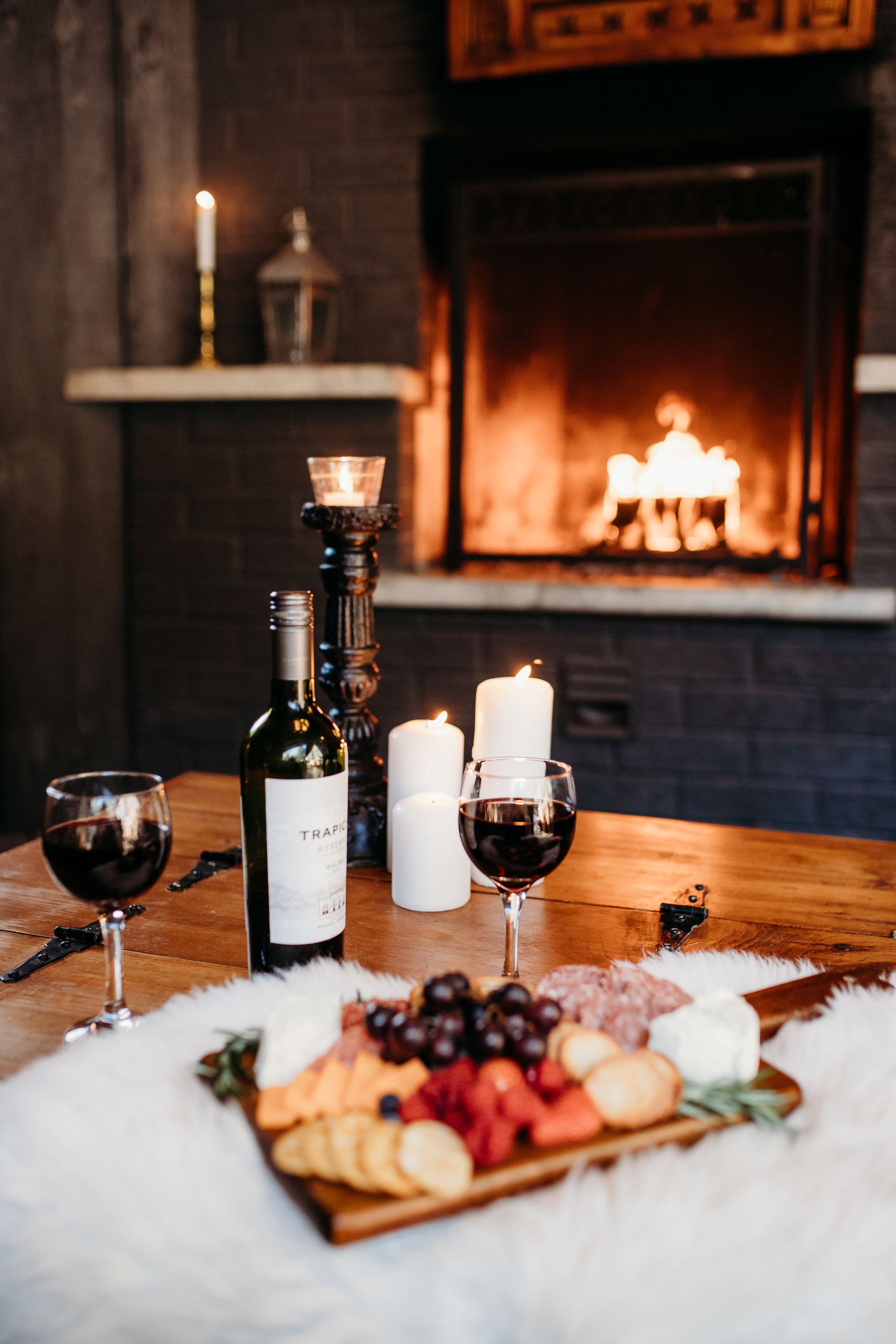 Wine, charcuterie board, and candles on a table in front of a fireplace.