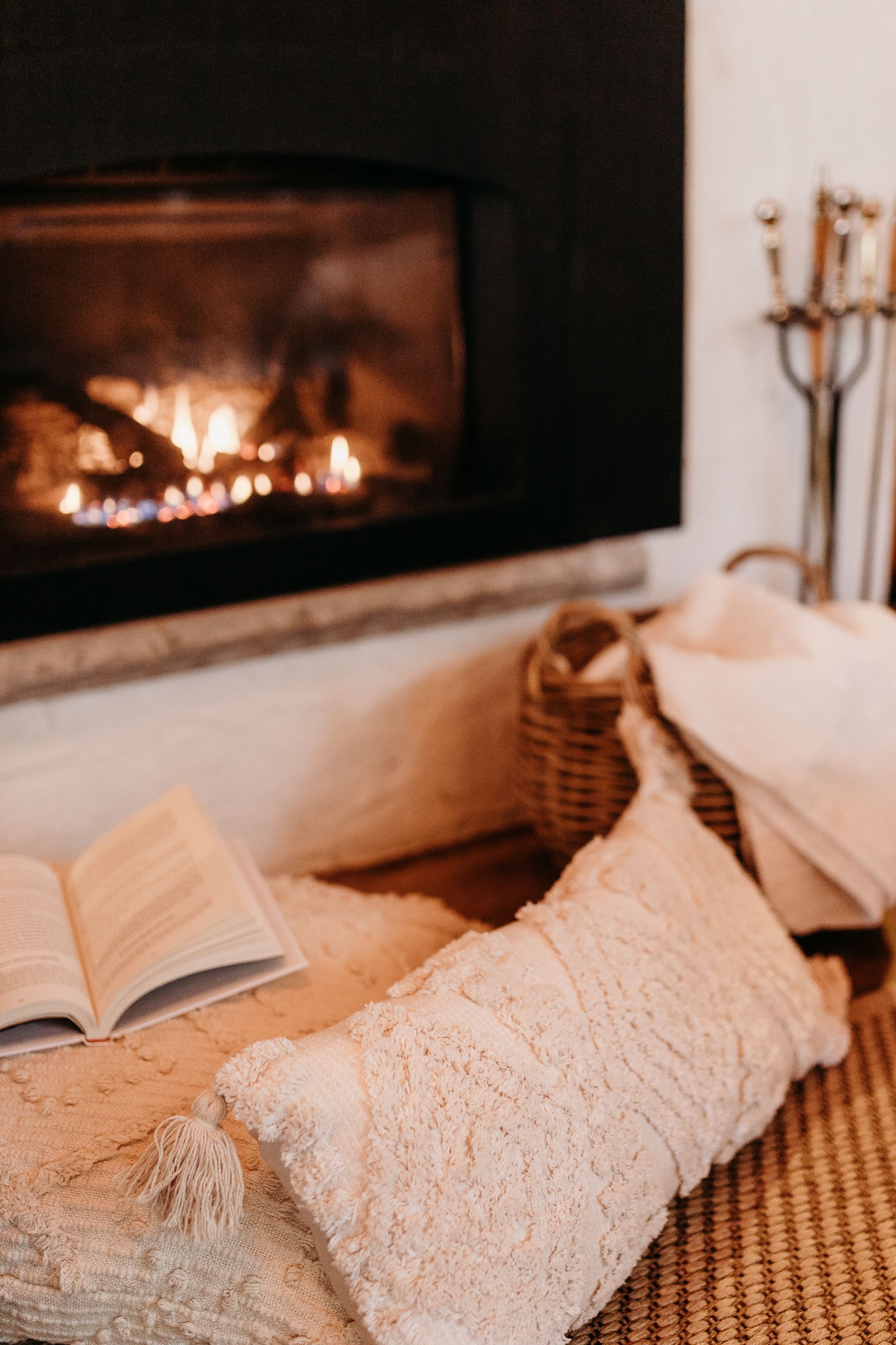 Fireplace with burning fire, open book, pillows, rug, and basket in warm setting.