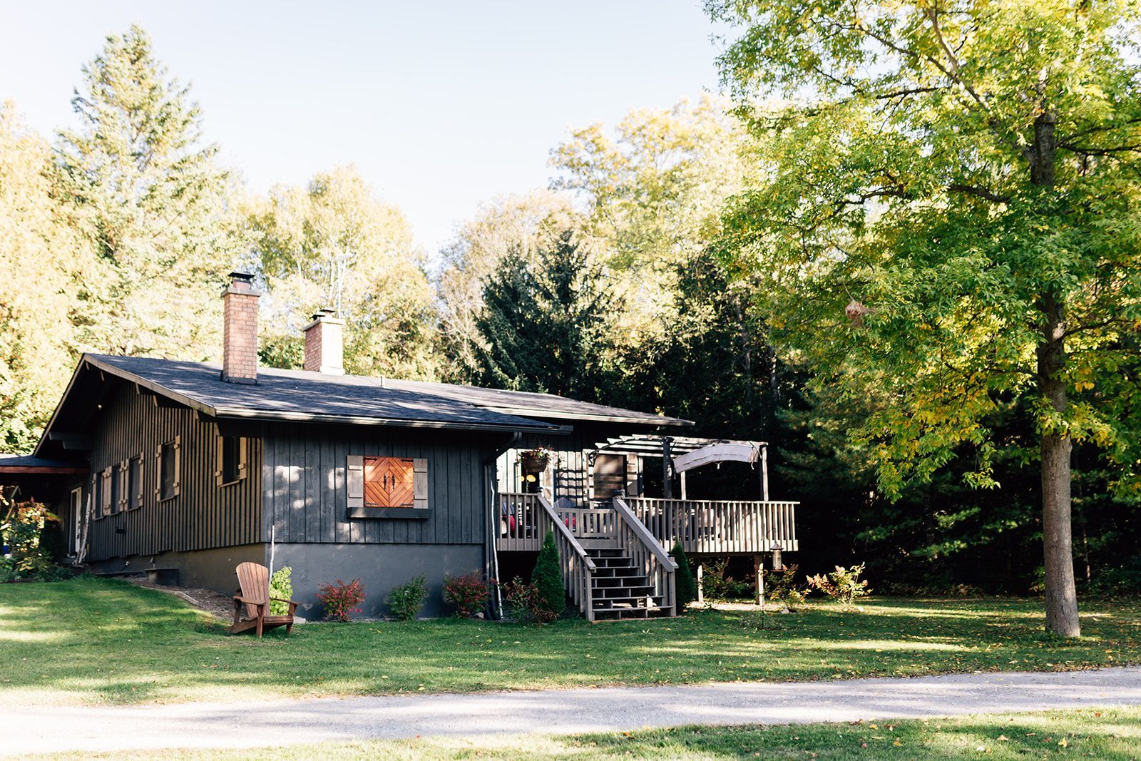 A dark grey cabin with a deck, chimney, and surrounding trees in a grassy yard.
