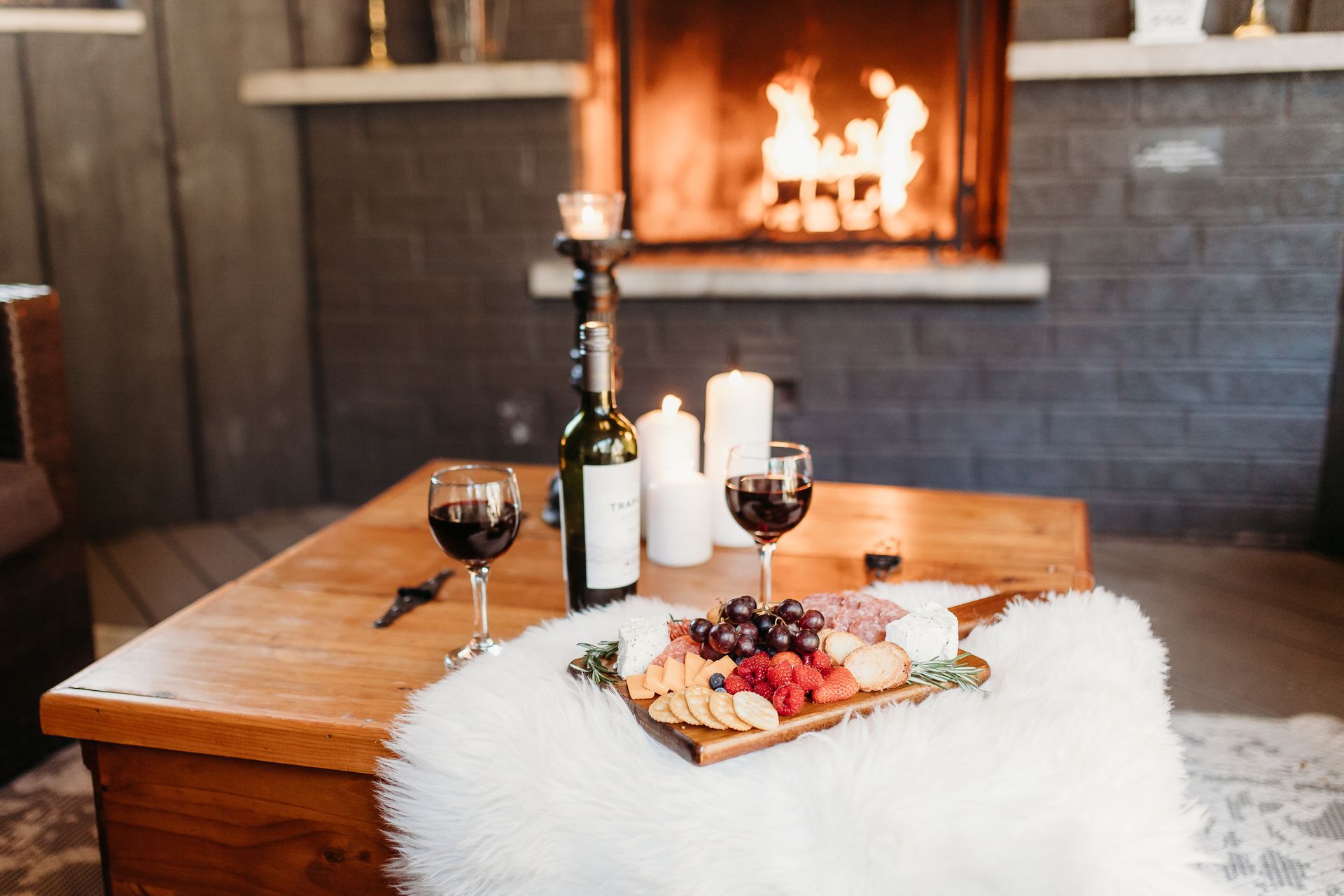Wine and charcuterie board on a wooden table in front of a fireplace with candles.
