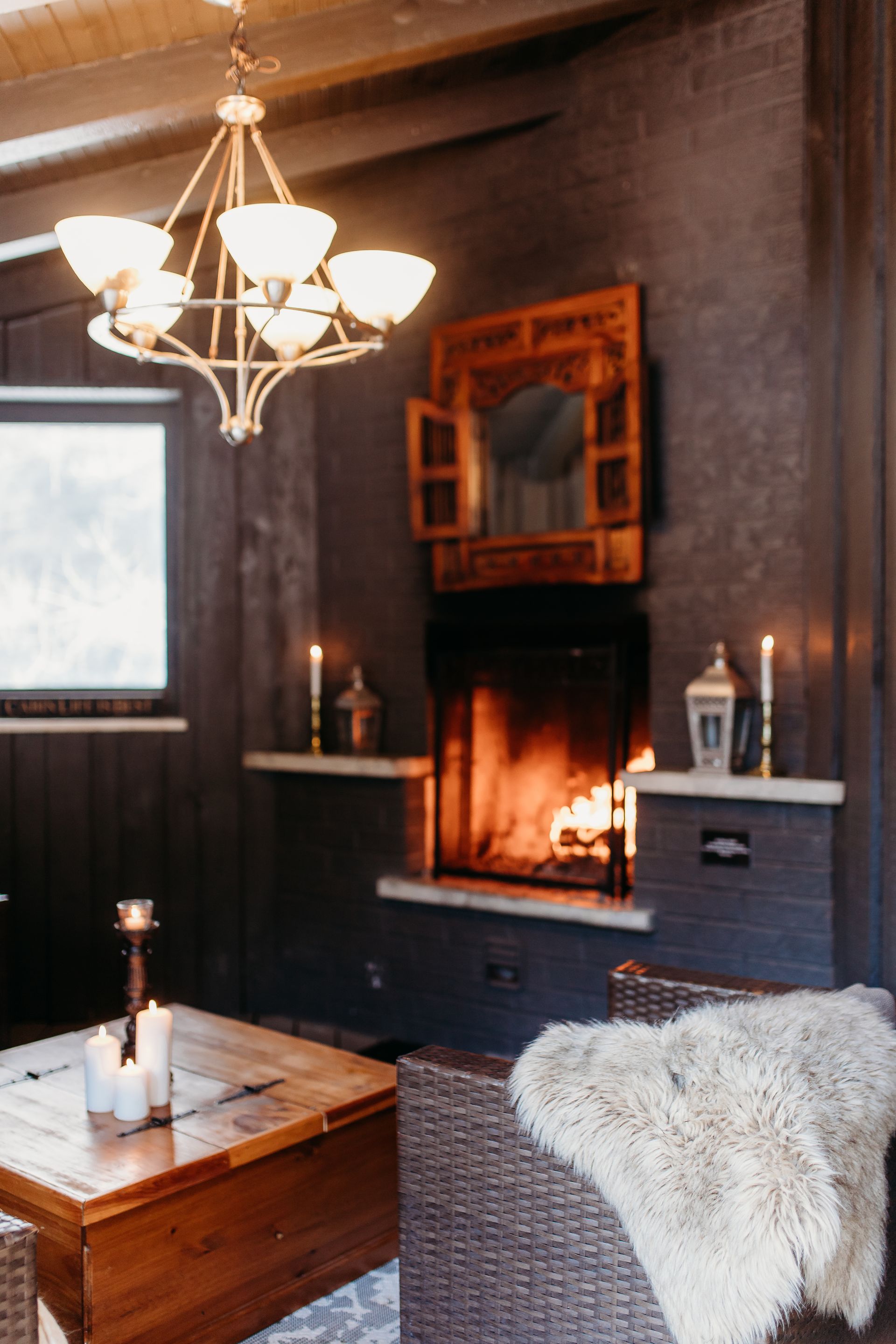 Cozy living room with fireplace, candles, and chandelier. Wooden walls, a rug, and a furry blanket create a warm atmosphere.