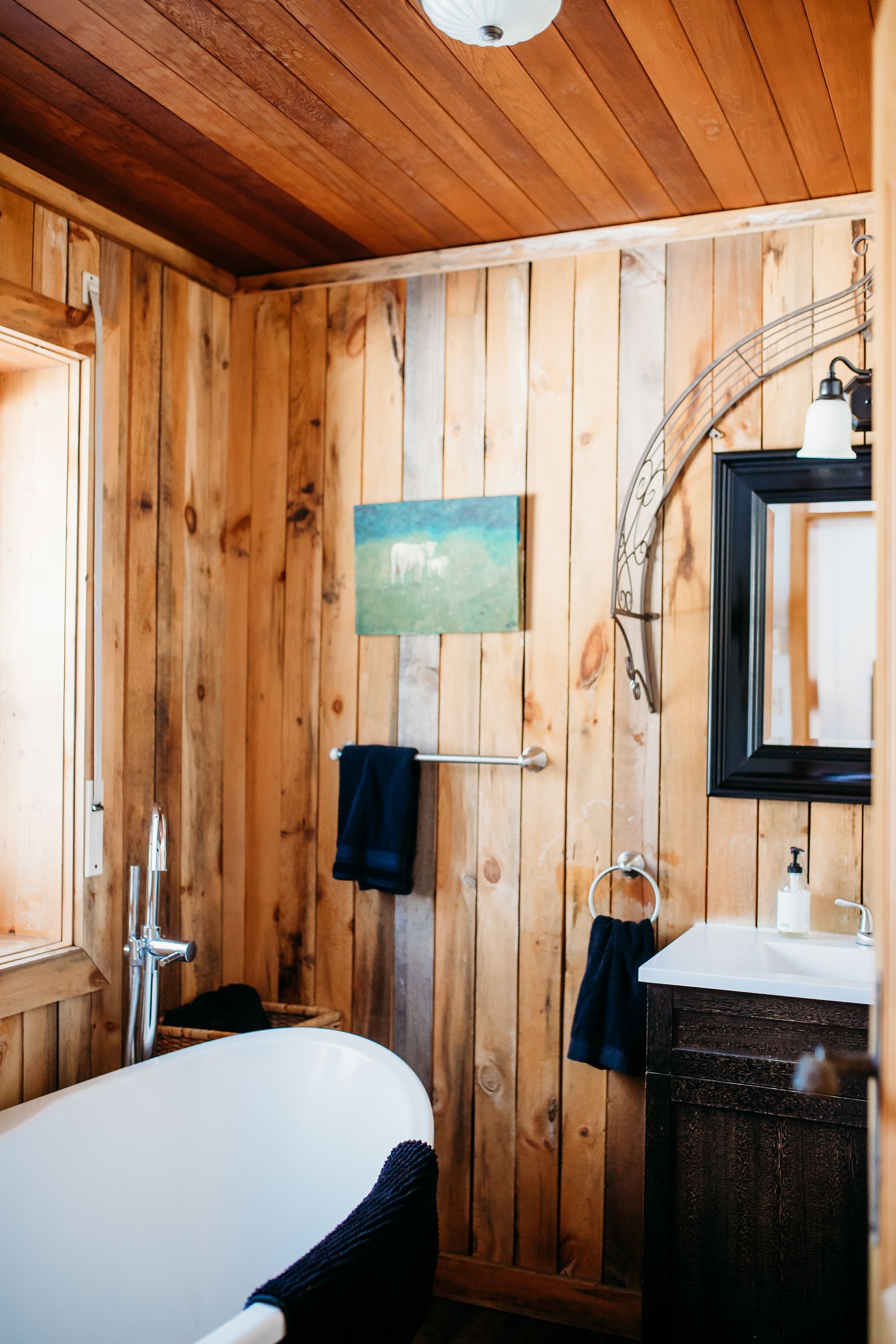 Rustic bathroom with wooden walls, white bathtub, black vanity, and artwork.