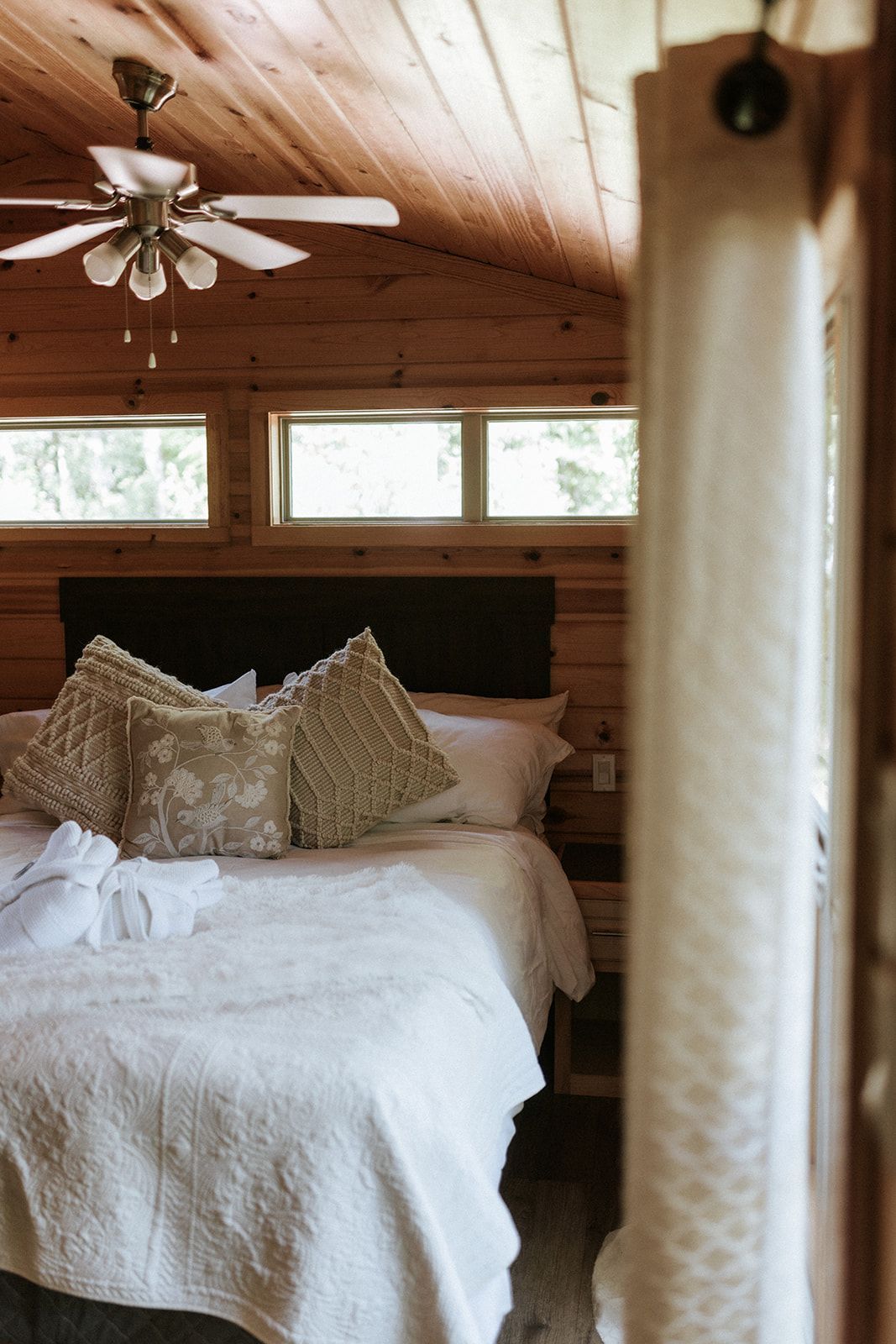 Cozy bedroom with white bedding, decorative pillows, wood paneling, windows, and a ceiling fan.