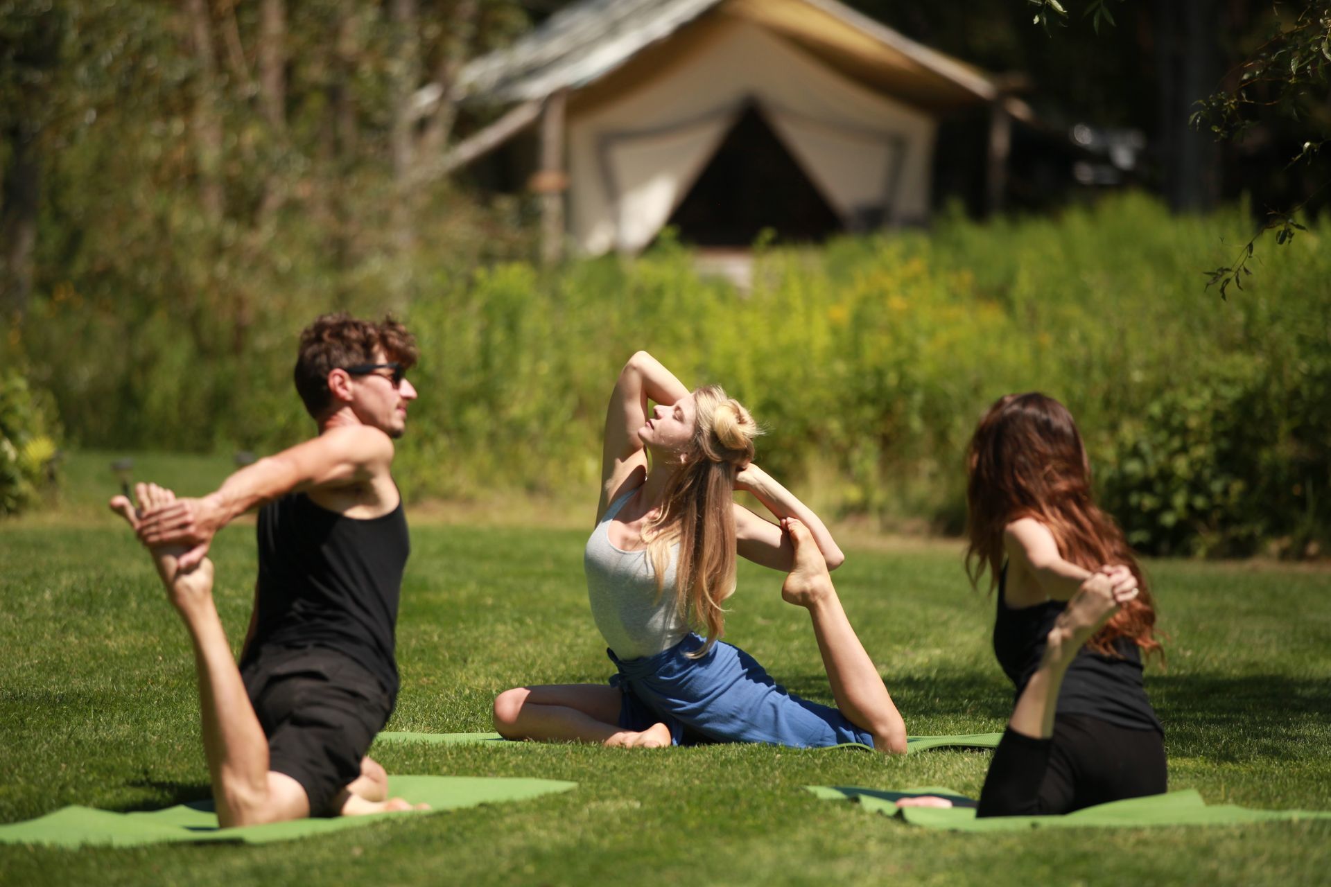 Three people in a park, doing yoga on mats. A tent is in the background.
