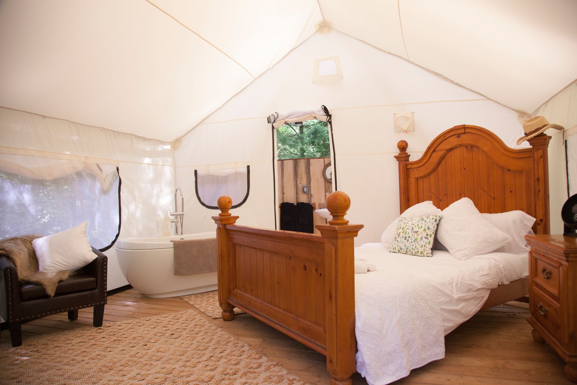 Interior of a glamping tent with a bed, bathtub, and chair, all in light colors and wood.