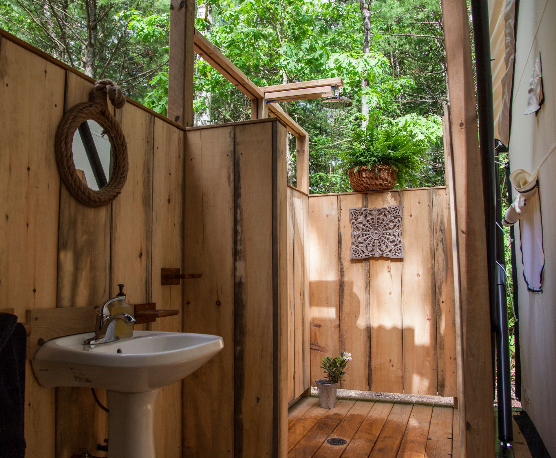 Rustic outdoor shower with wooden walls, sink, mirror, and open roof, surrounded by trees.