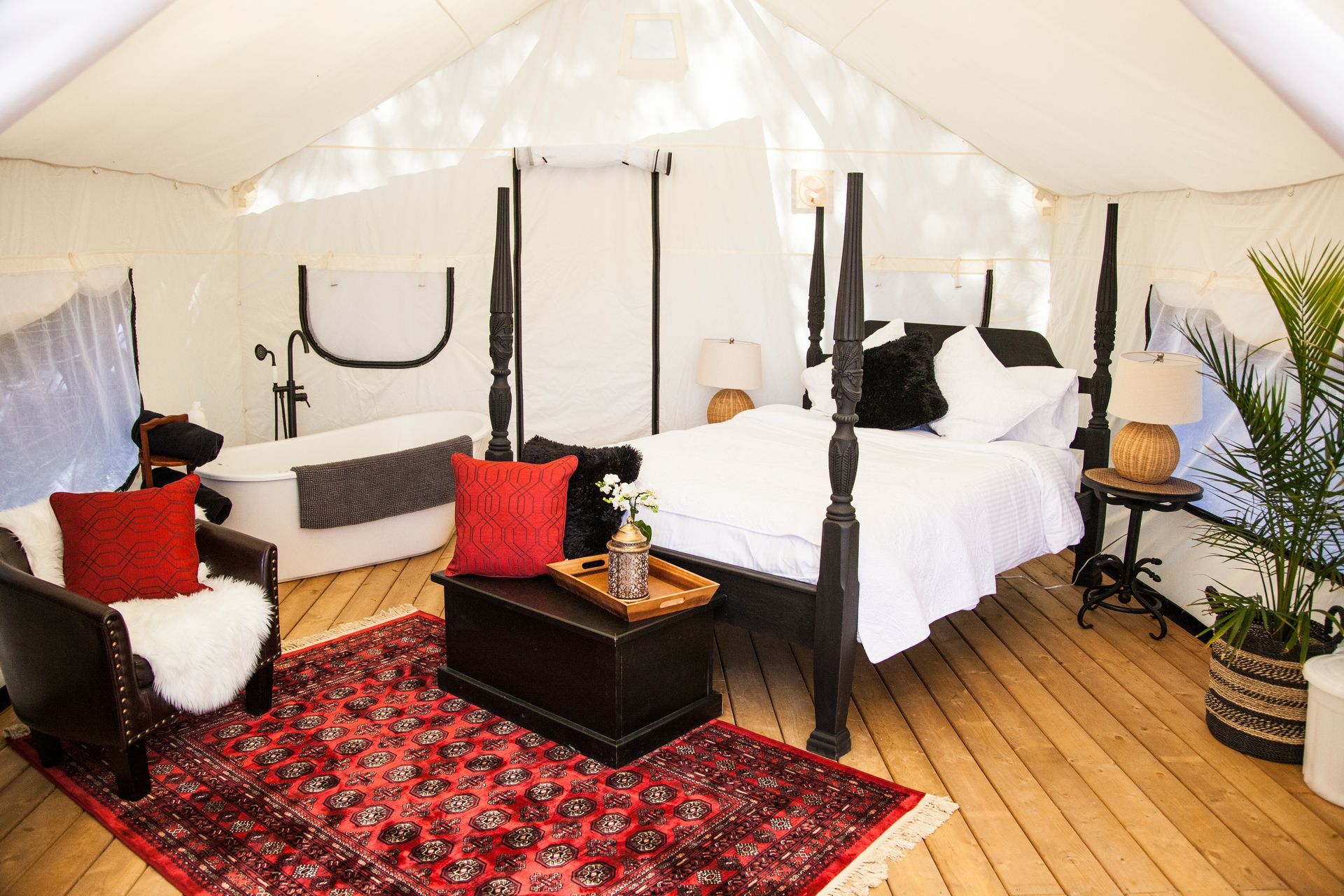 A luxurious tent interior with a four-poster bed, bathtub, sitting area, and plant on a wooden floor.