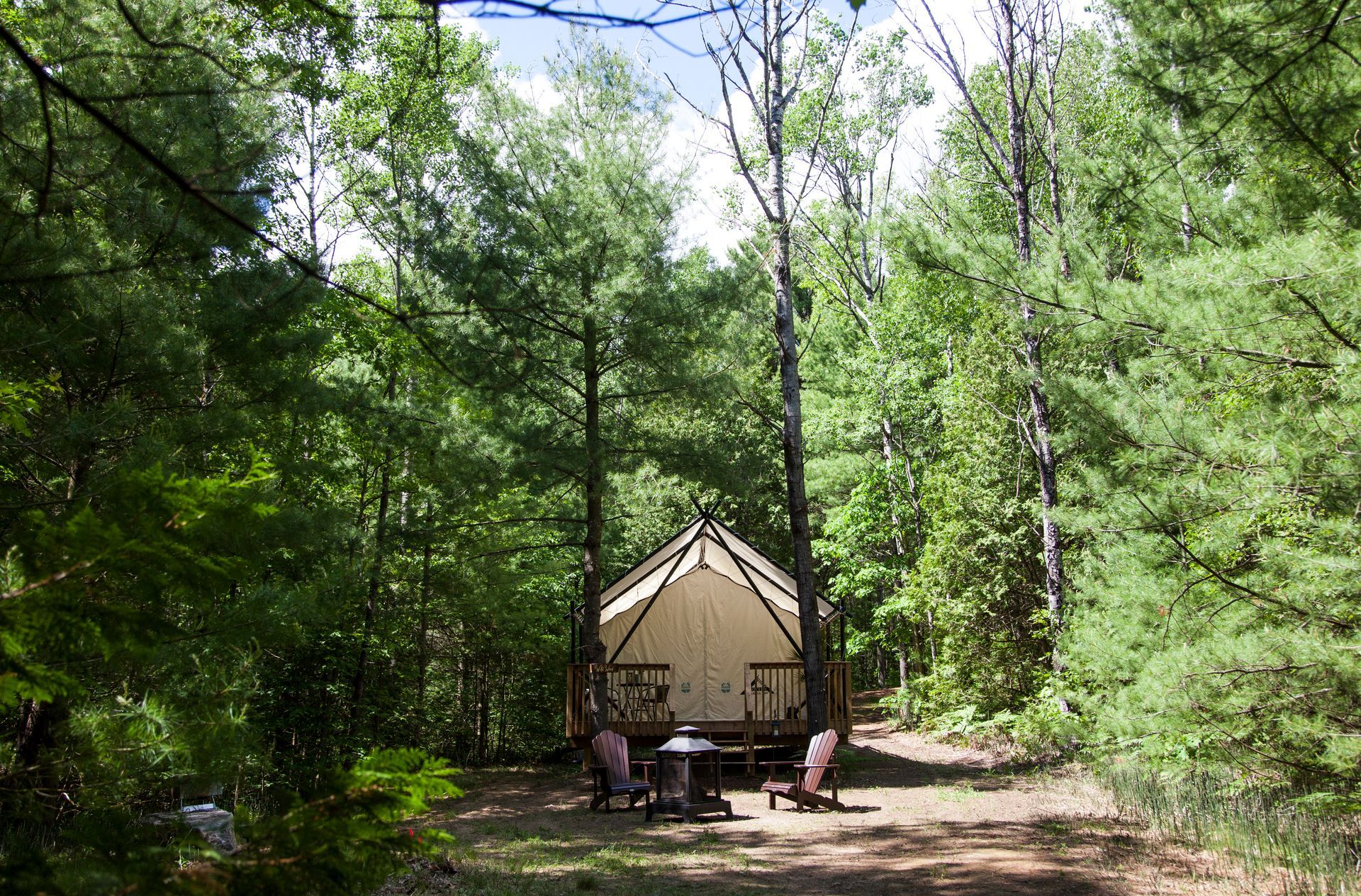 Canvas tent set up in a clearing in the woods, with trees and chairs around a fire pit.