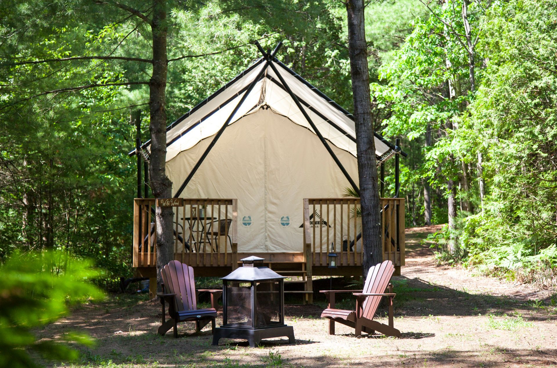 Glamping tent on a wooden deck in a wooded area with chairs and a fire pit.