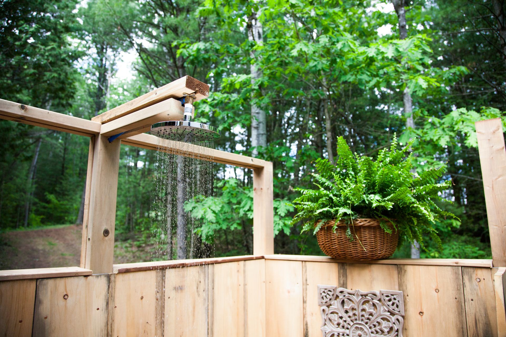 Outdoor wooden shower with water falling; fern in a hanging basket, trees in background.