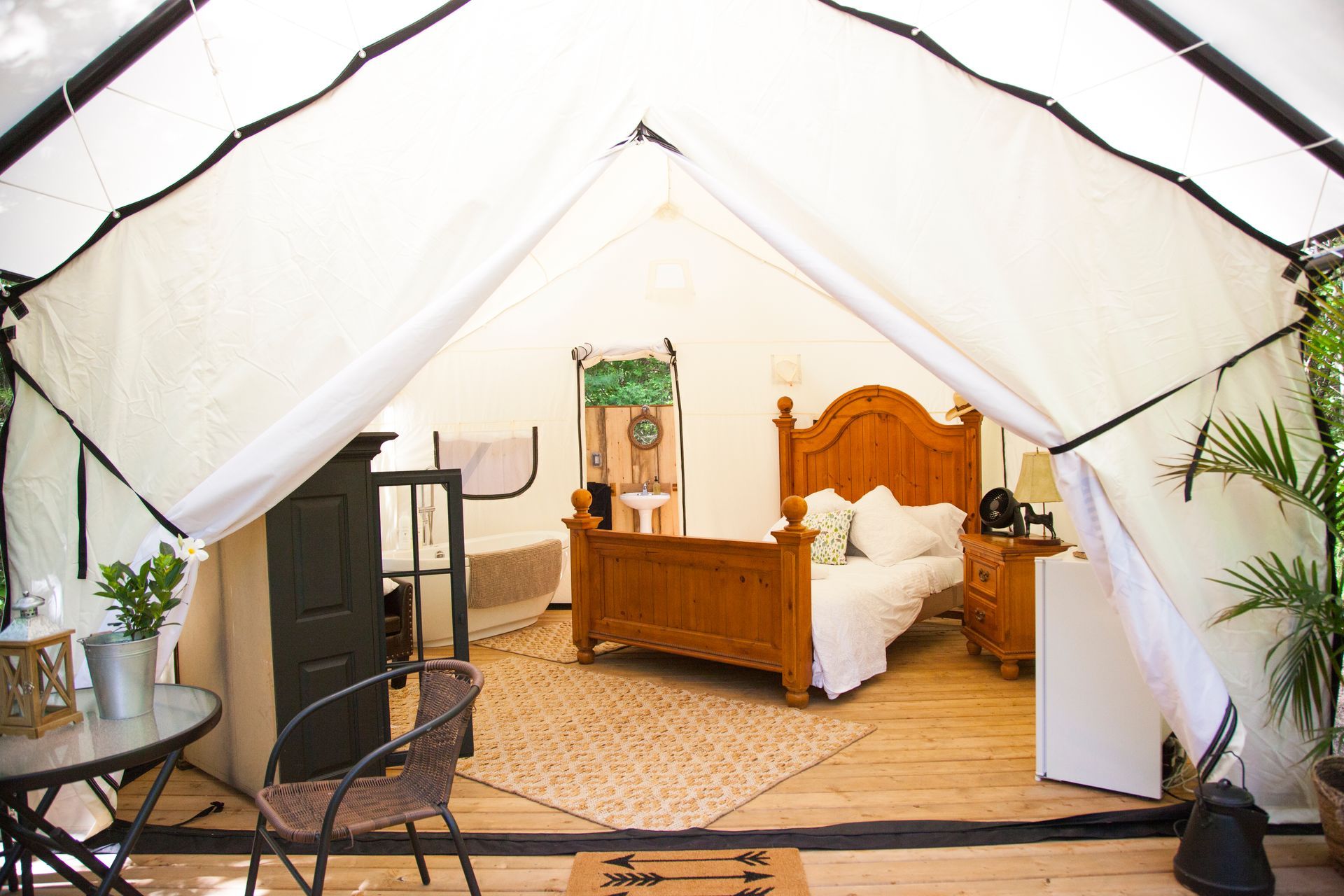 Inside a glamping tent: a wooden bed, nightstand, and rug. A table and chair are outside.